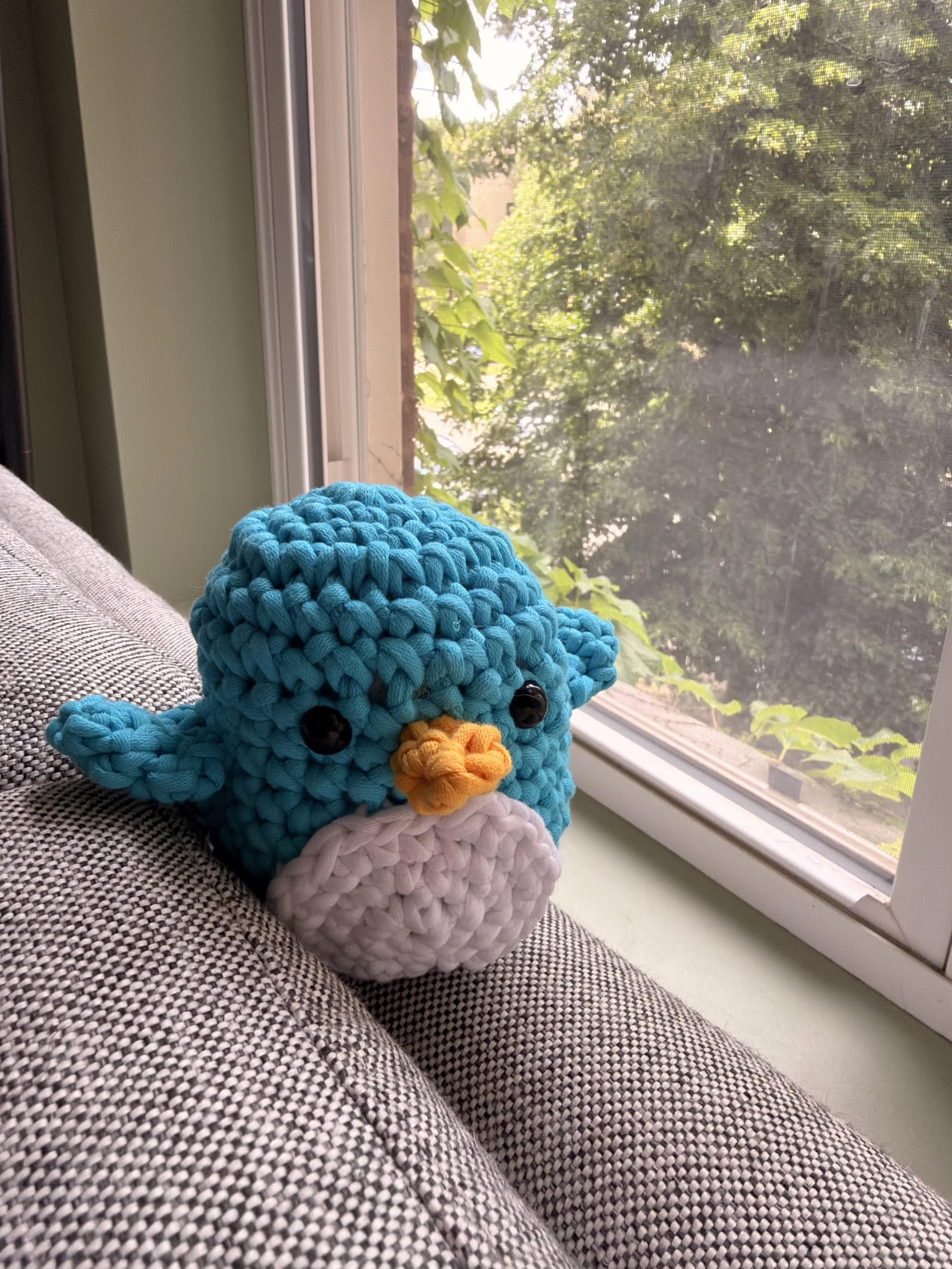 A knitted blue bird plush toy with a yellow beak and black eyes sitting on a gray fabric surface near a window with green foliage outside.