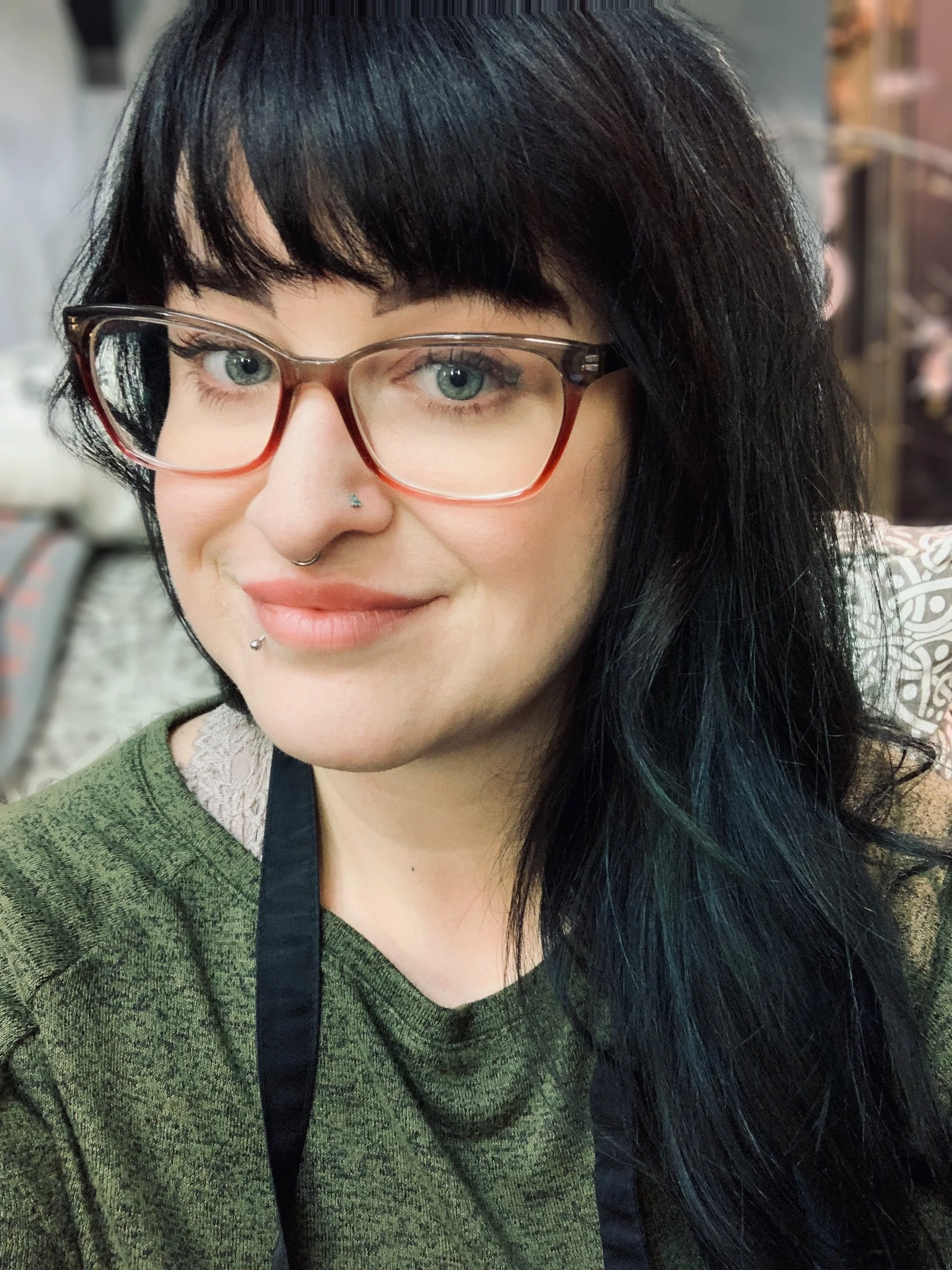 Close-up of a person with black hair, light skin, wearing glasses, with a septum piercing, and a smile, in an indoor setting.