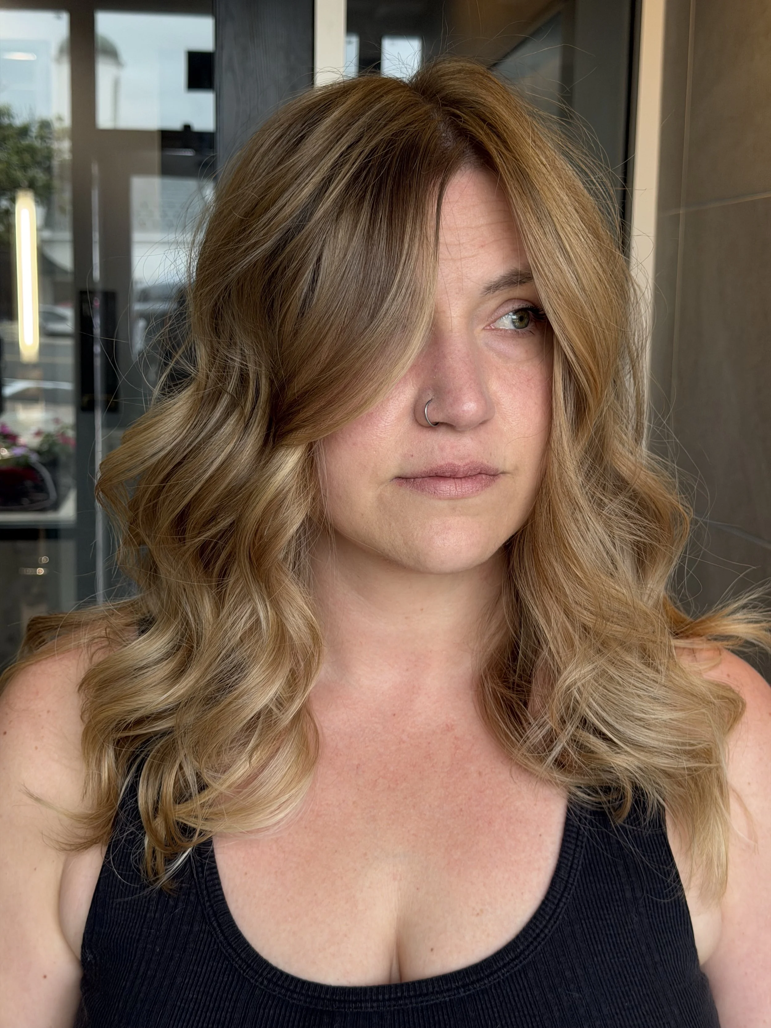 A woman with wavy blonde hair and green eyes wearing a black tank top, visible from her shoulders up, inside a building with large windows and outdoor scene in the background.