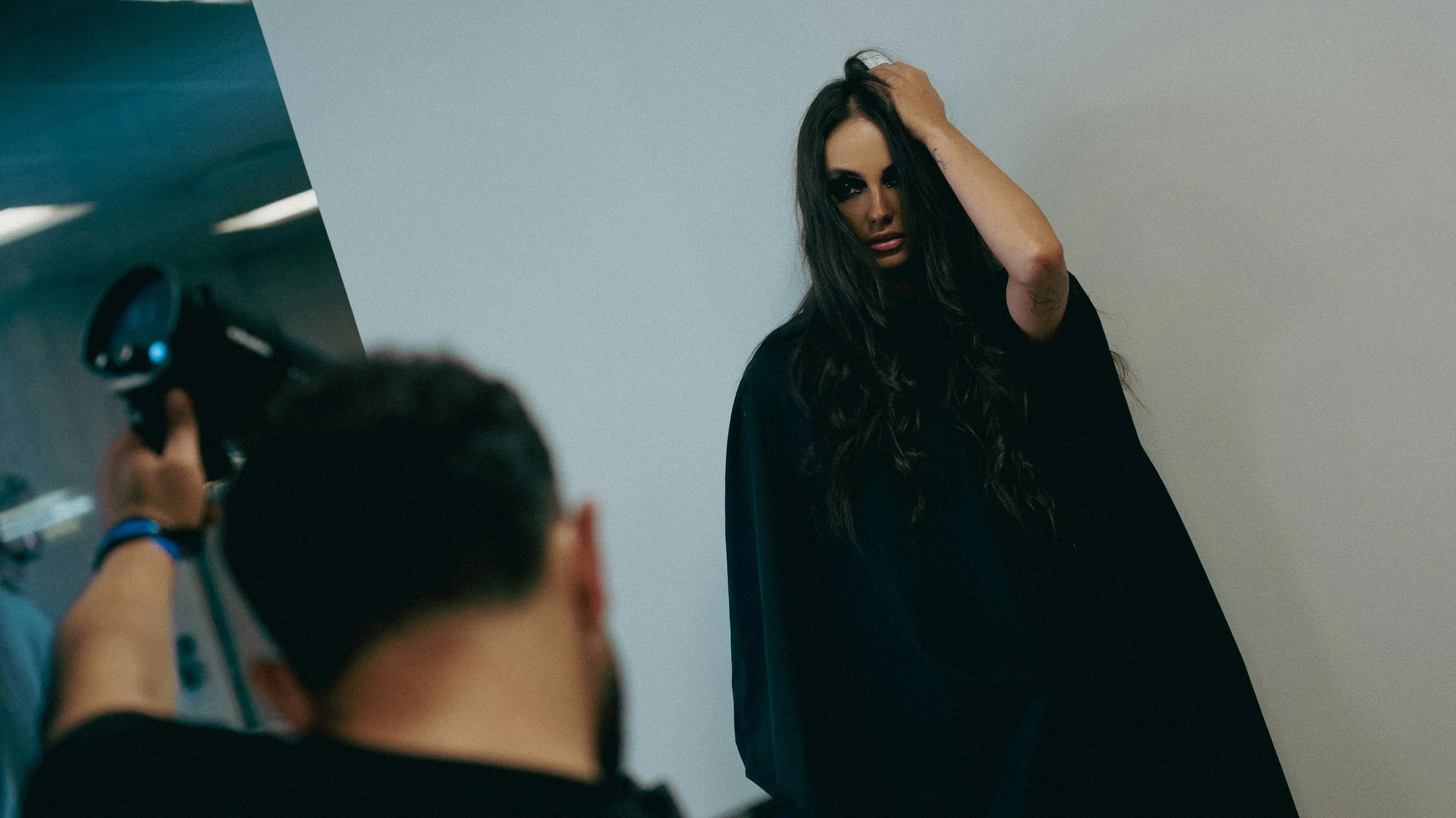 A woman with long dark hair and dark eye makeup posing for a photo at a photoshoot, with a photographer taking her picture.