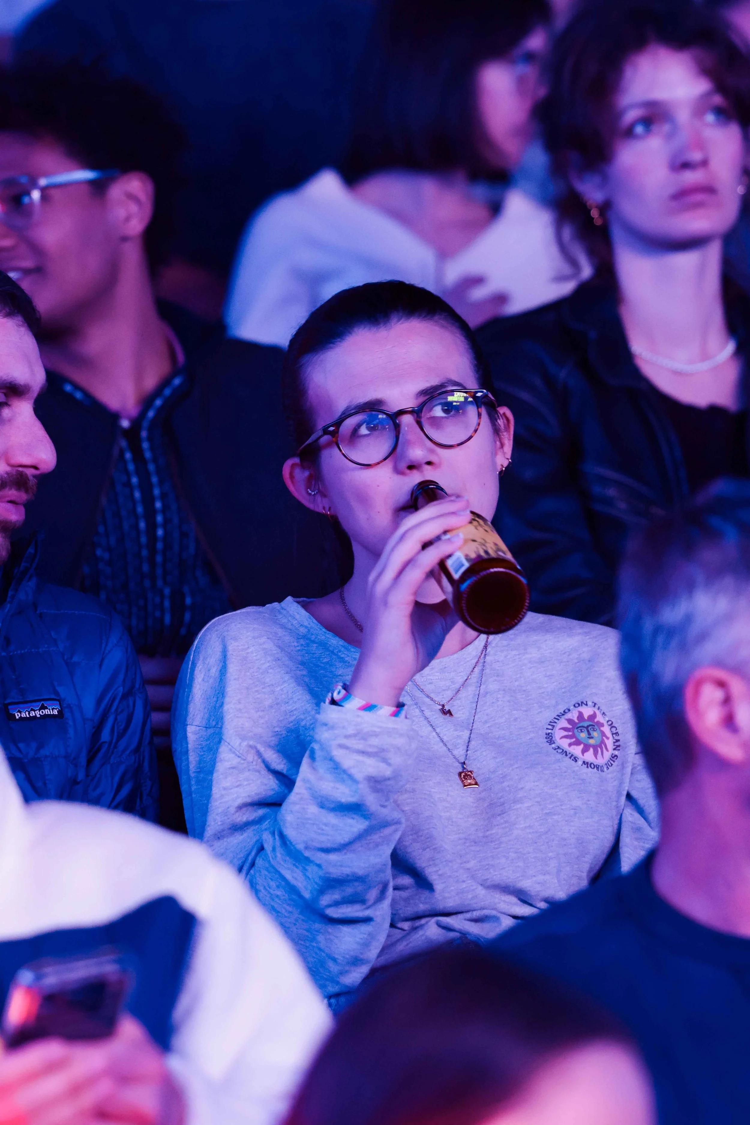 Public assistant à un concert, baigné de lumières bleues et violettes, avec une personne au premier plan portant des lunettes et tenant une canette (événement La Nuit du Trail).