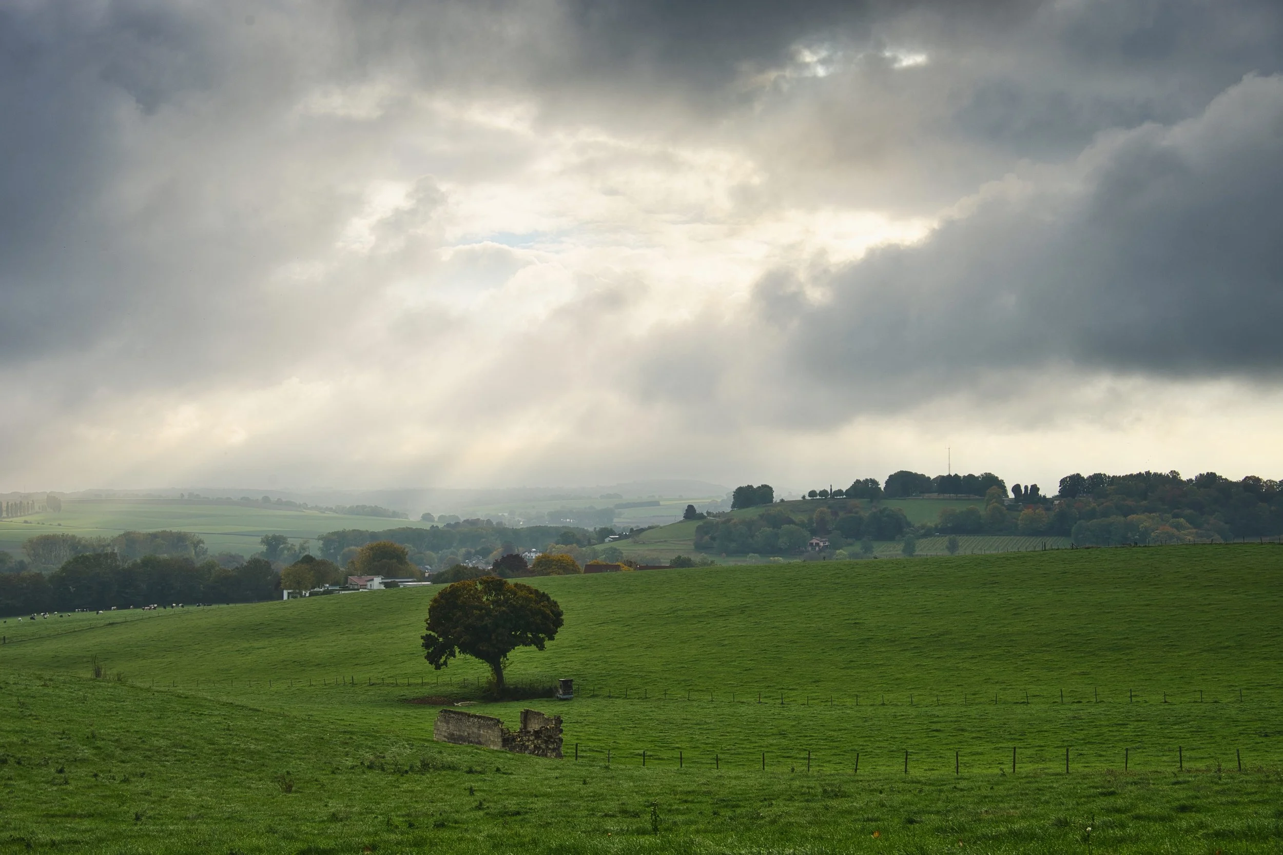Een uitgestrekt groen landschap met enkele bomen, boerderijen en een heuvelachtig terrein onder een bewolkte hemel met donkere wolken.