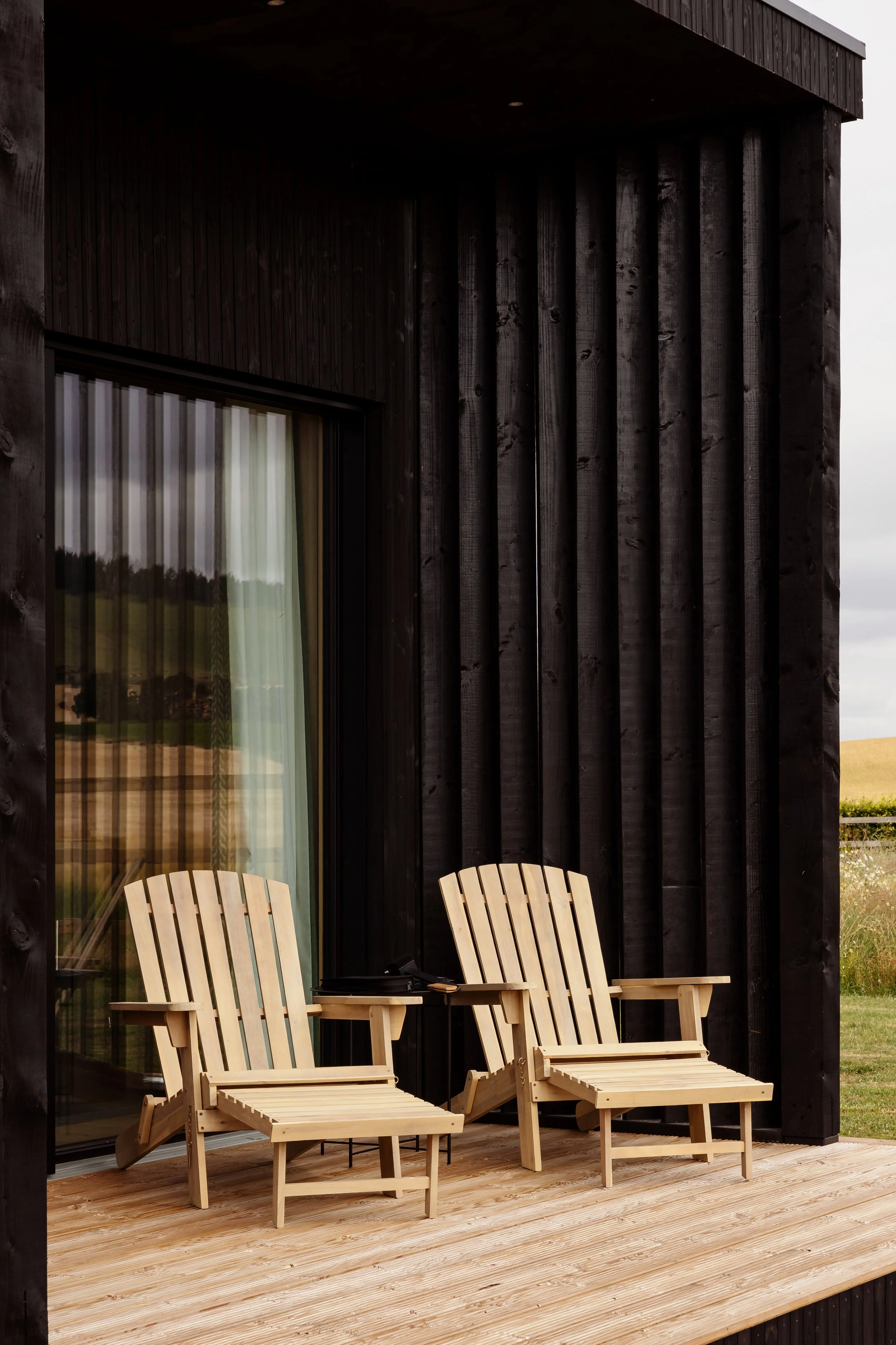 A wooden deck with two wooden chairs, located outside a modern black cabin with large glass sliding doors.