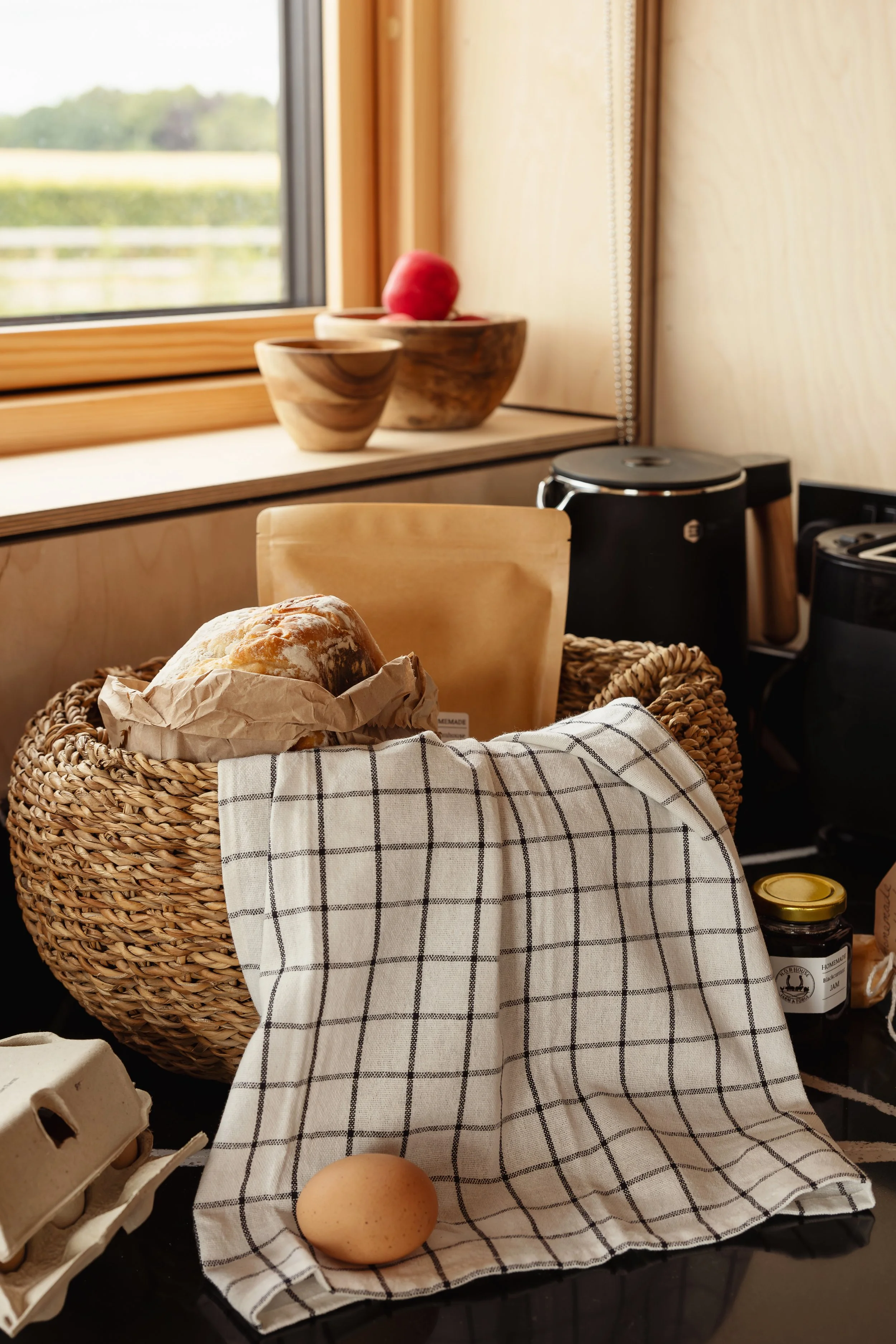 A kitchen counter with a woven basket of bread, a white and black checkered cloth, two eggs, and various kitchen appliances near a window.