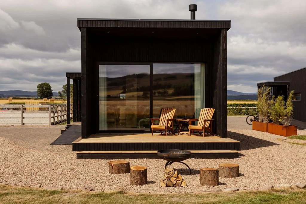 Black timber single storey cabin. Large sliding doors. Fire pit area with gravel.