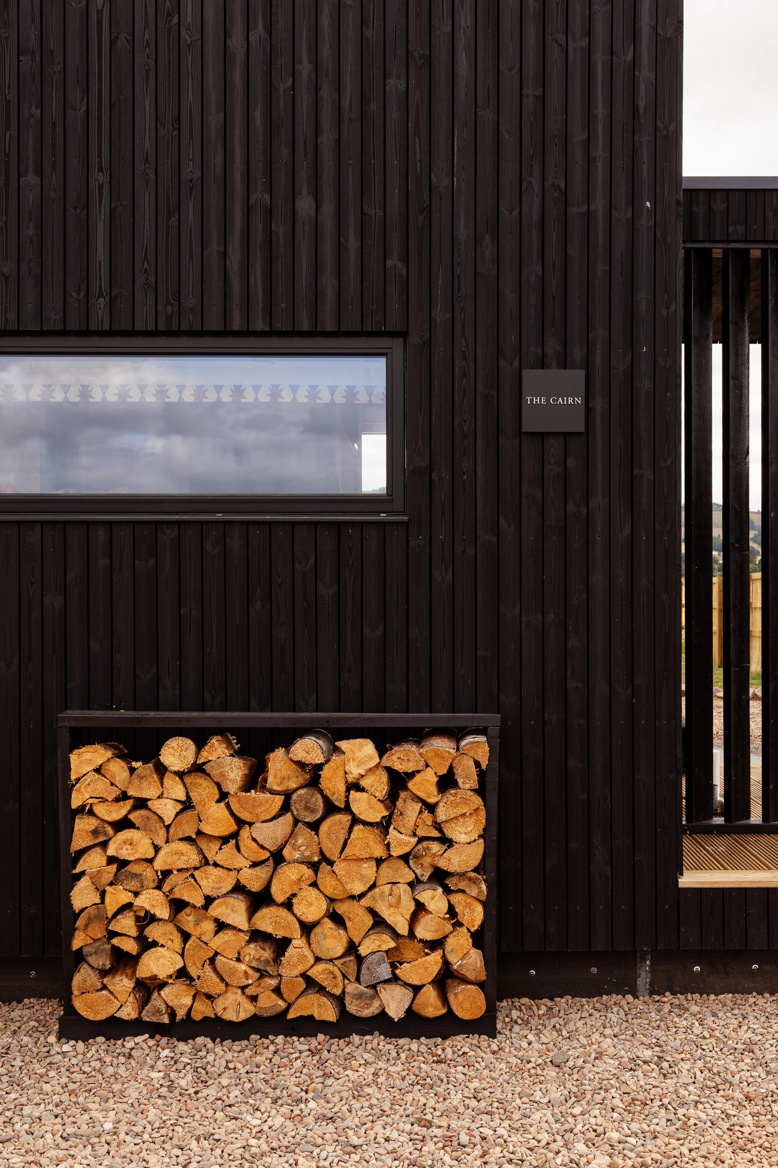 Wooden exterior wall with a horizontal window, a black sign reading 'The Cairn', a firewood storage area with logs, and gravel ground.