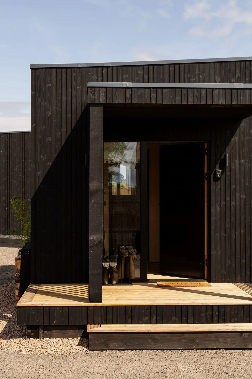 Black timber cabin with porch area. Decked. Wellie rack. Door half open showing entrance wall.