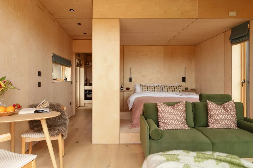 Open plan bedroom, living, dining area. Velvet green sofa and pink cushions. Layered bedding. 