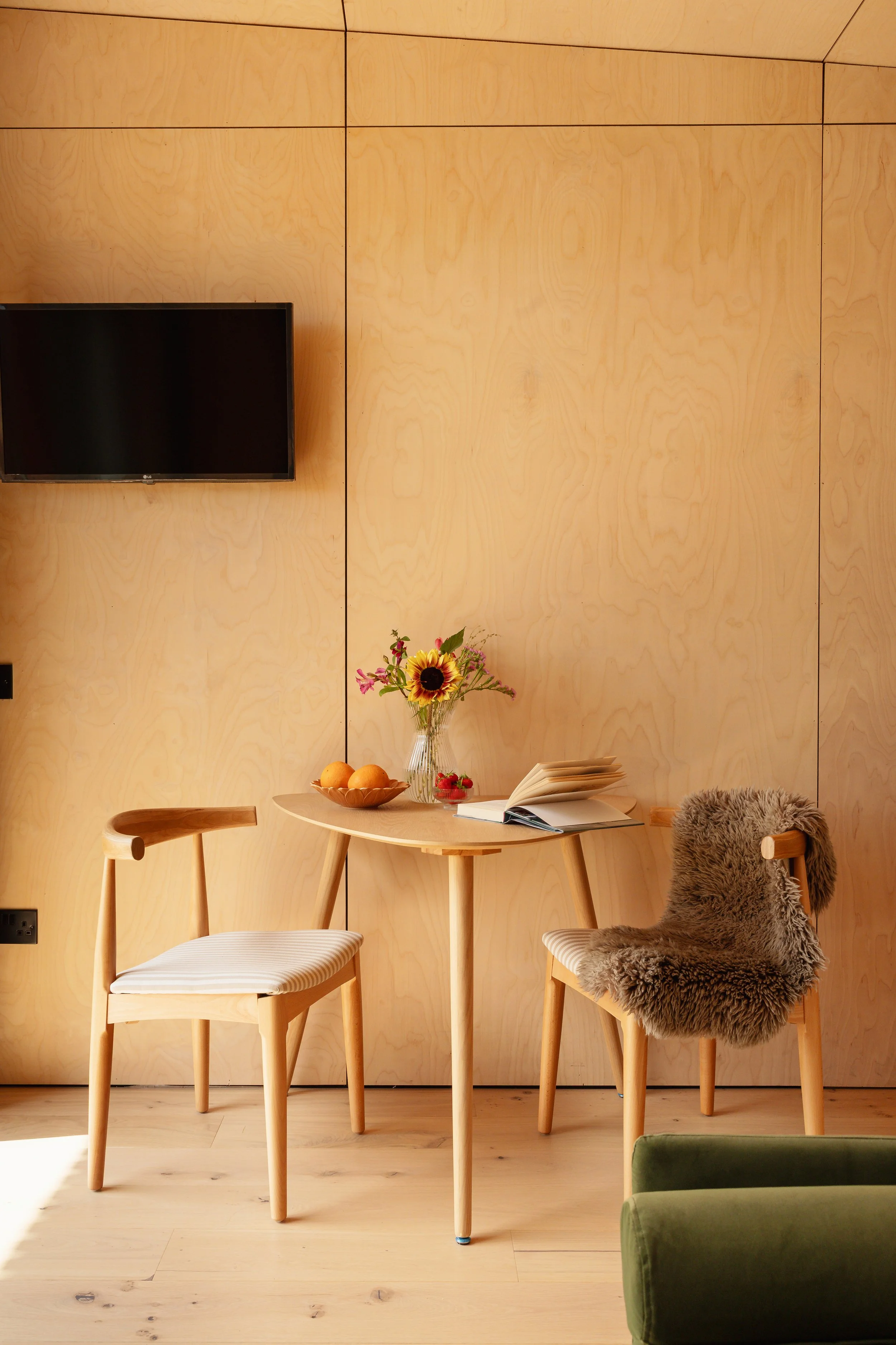 Interior of a cosy cabin with a wooden dining table, two chairs, one with a furry throw, a vase with sunflowers, a bowl of fruit, an open book, and a wall-mounted flat-screen TV.