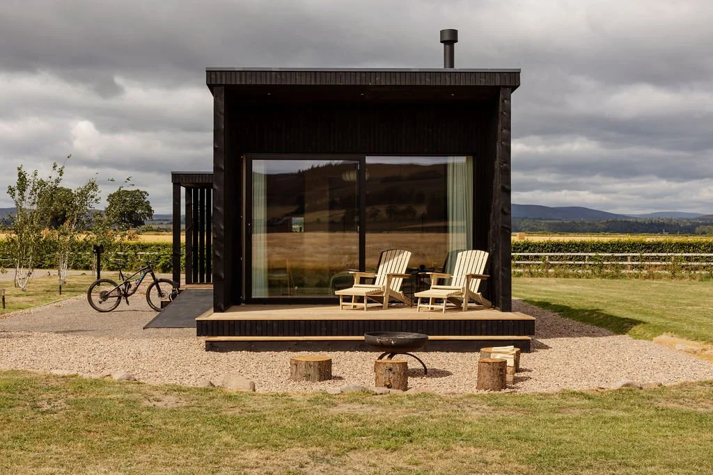 Black timber cabin with overhang. Decked area with sun loungers. Bike in mounted bike rack. Disabled ramp access. Firepit area with rustic logs. Angus hill views beyond.