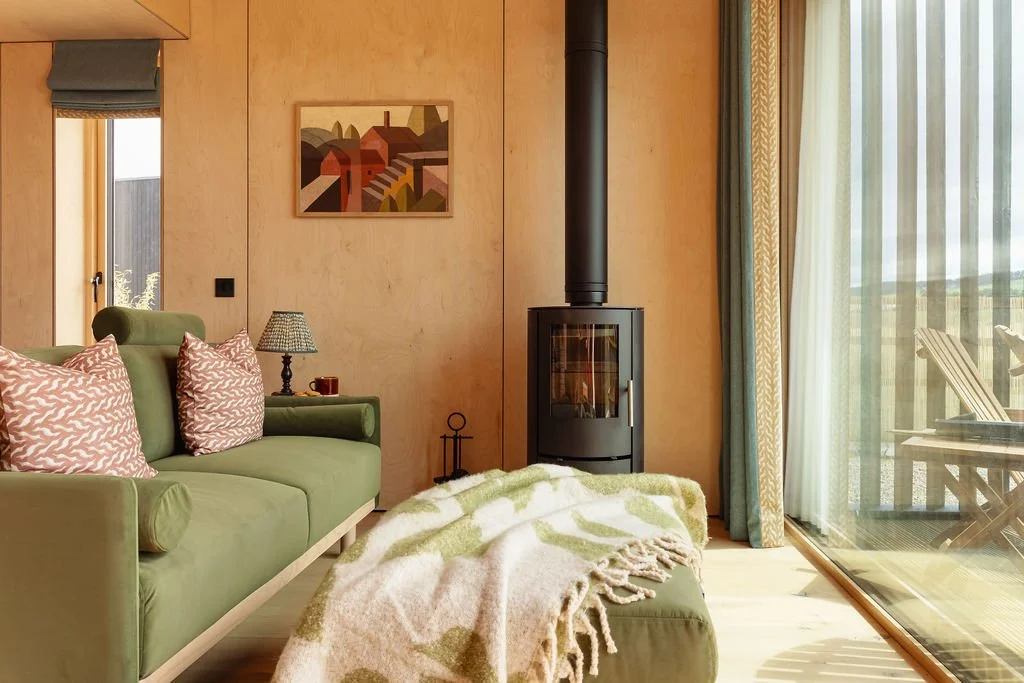 Scandi style living space. Green sofa and foot stool with blanket. Pink cushions. Timber floors. Log-burner. Pooky light.