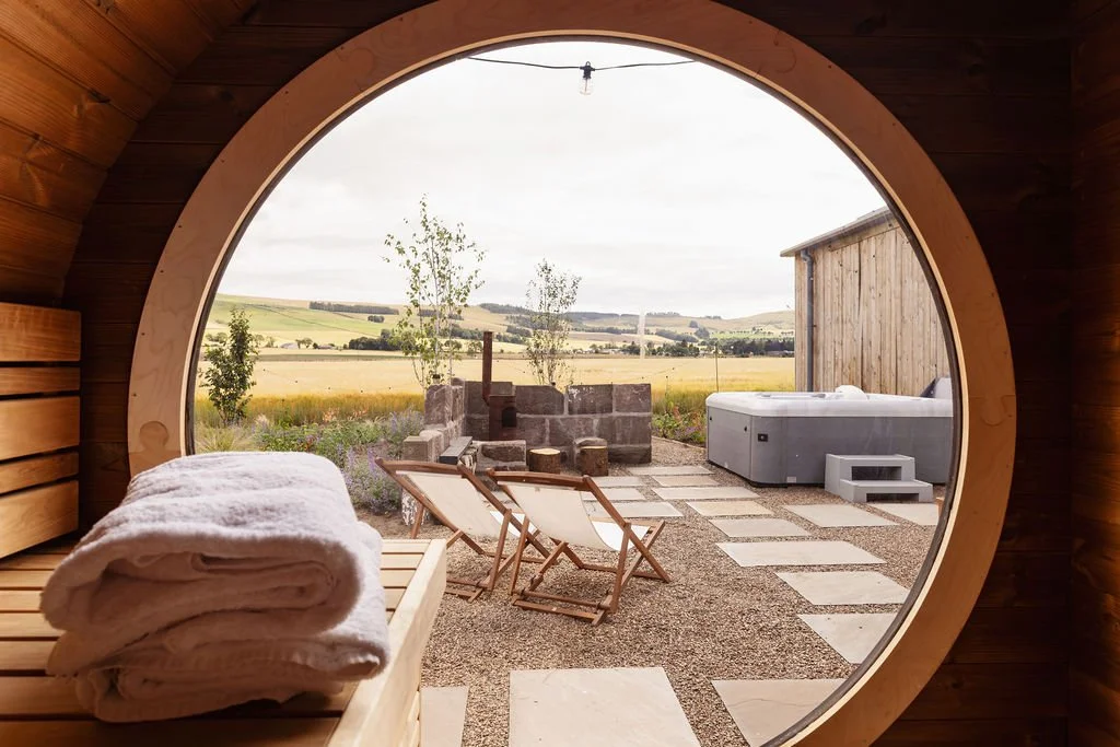 Sauna with round window overlooking a wellness area. Decked chairs, hot tub - walled firepit area. Farmland views.
