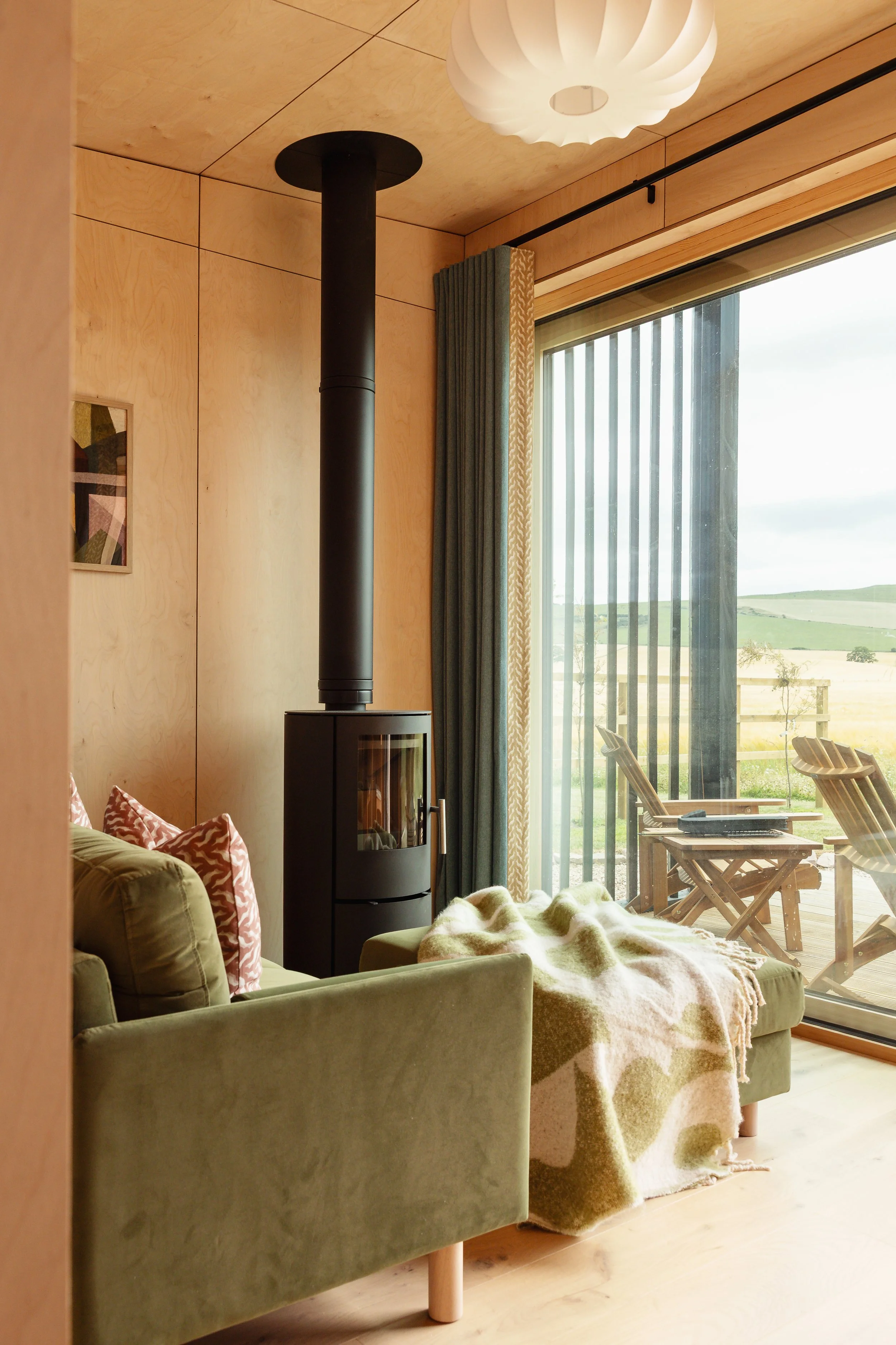 Living room with green velvet sofa, patterned cushions, wood stove, large window with curtains, and outdoor patio with wooden chairs and a view of rolling hills.