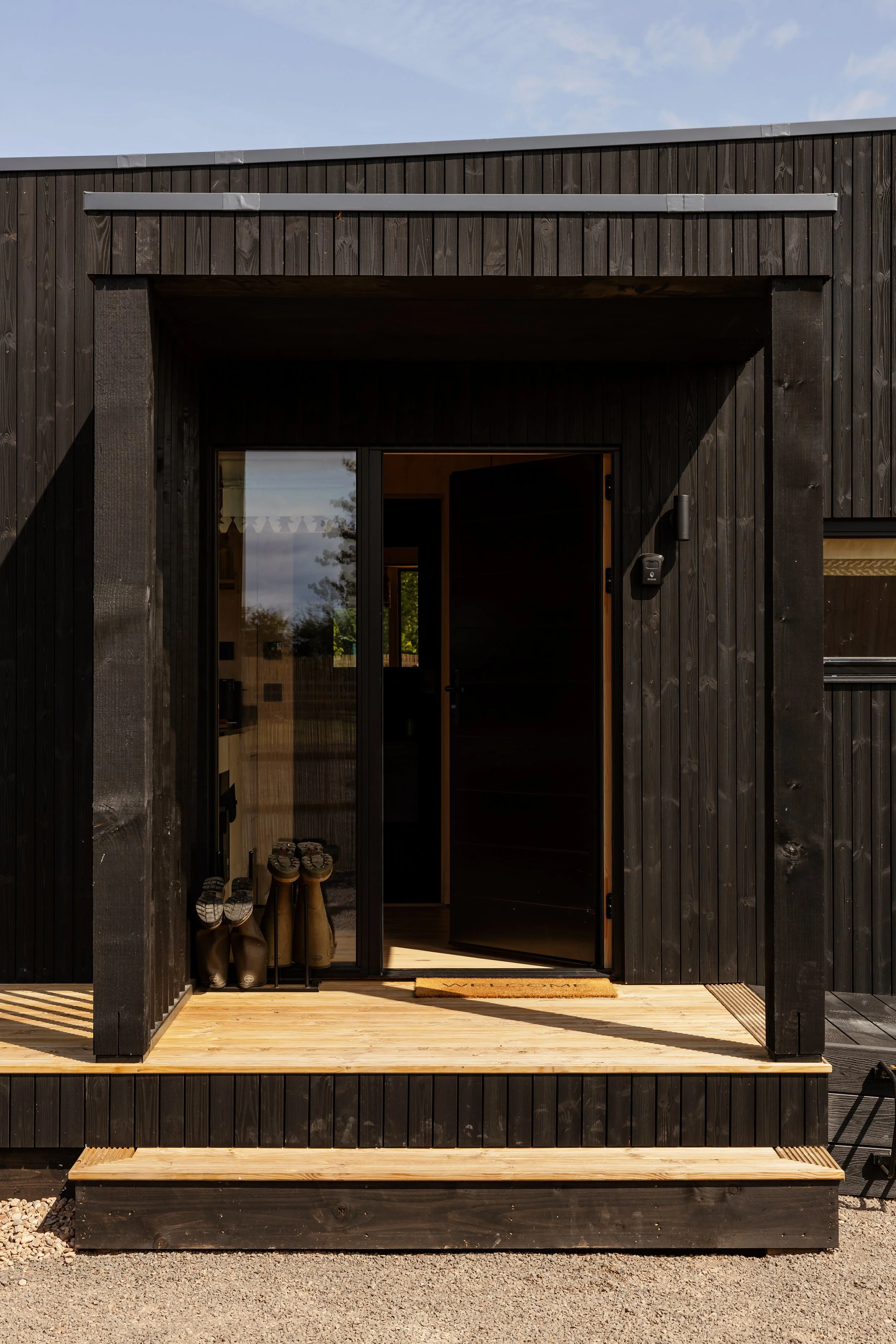 Front view of a black wooden house with an open glass door, showing shoes and boots inside. There is a doormat outside the door, and a small black outdoor chair on the right. The house has a wooden deck and a clear sky with some clouds above.