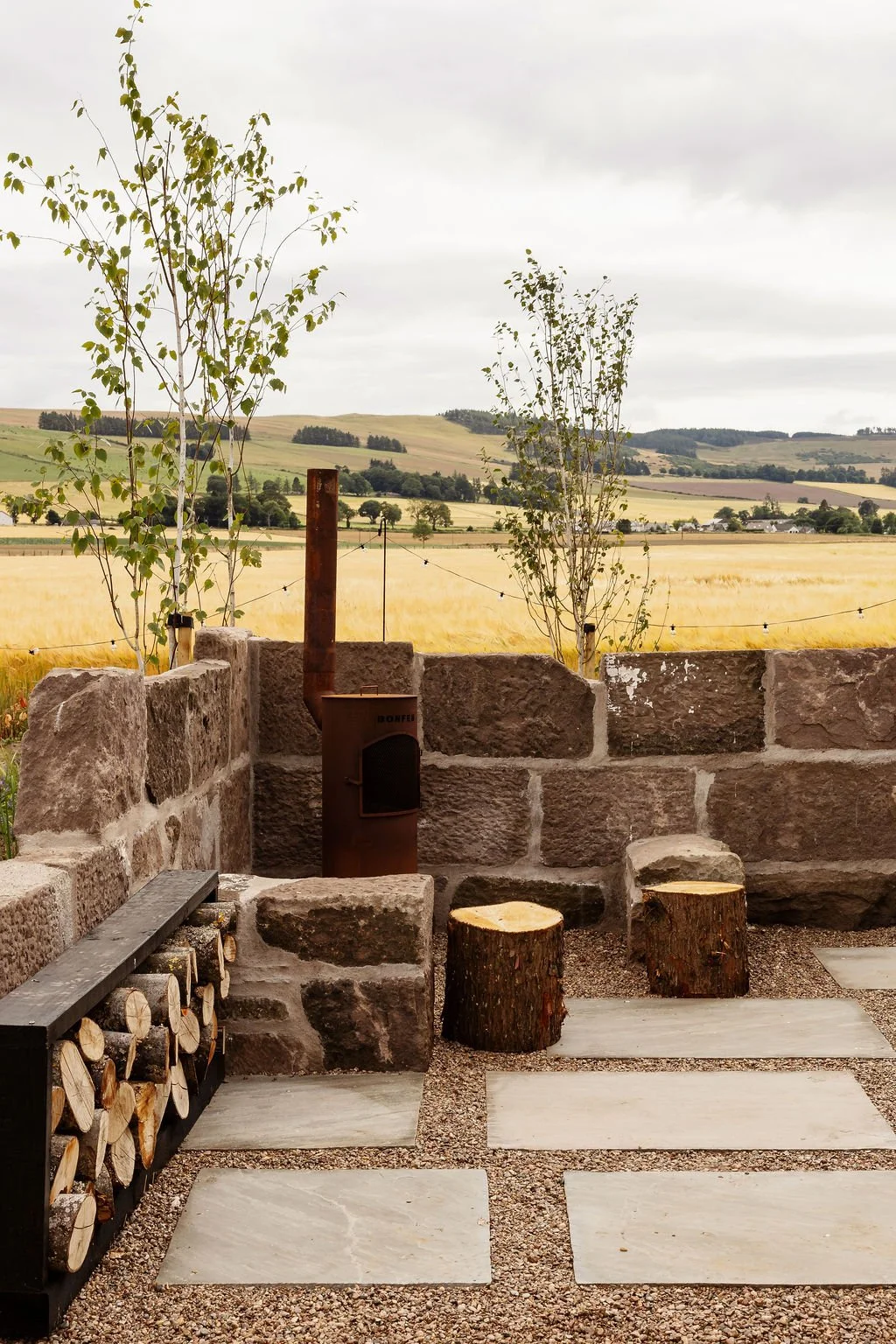 Walled fire pit area with stone setting and rustic logs. Stepping stones. Stack of logs. Two silver birch tress against farmland views. 