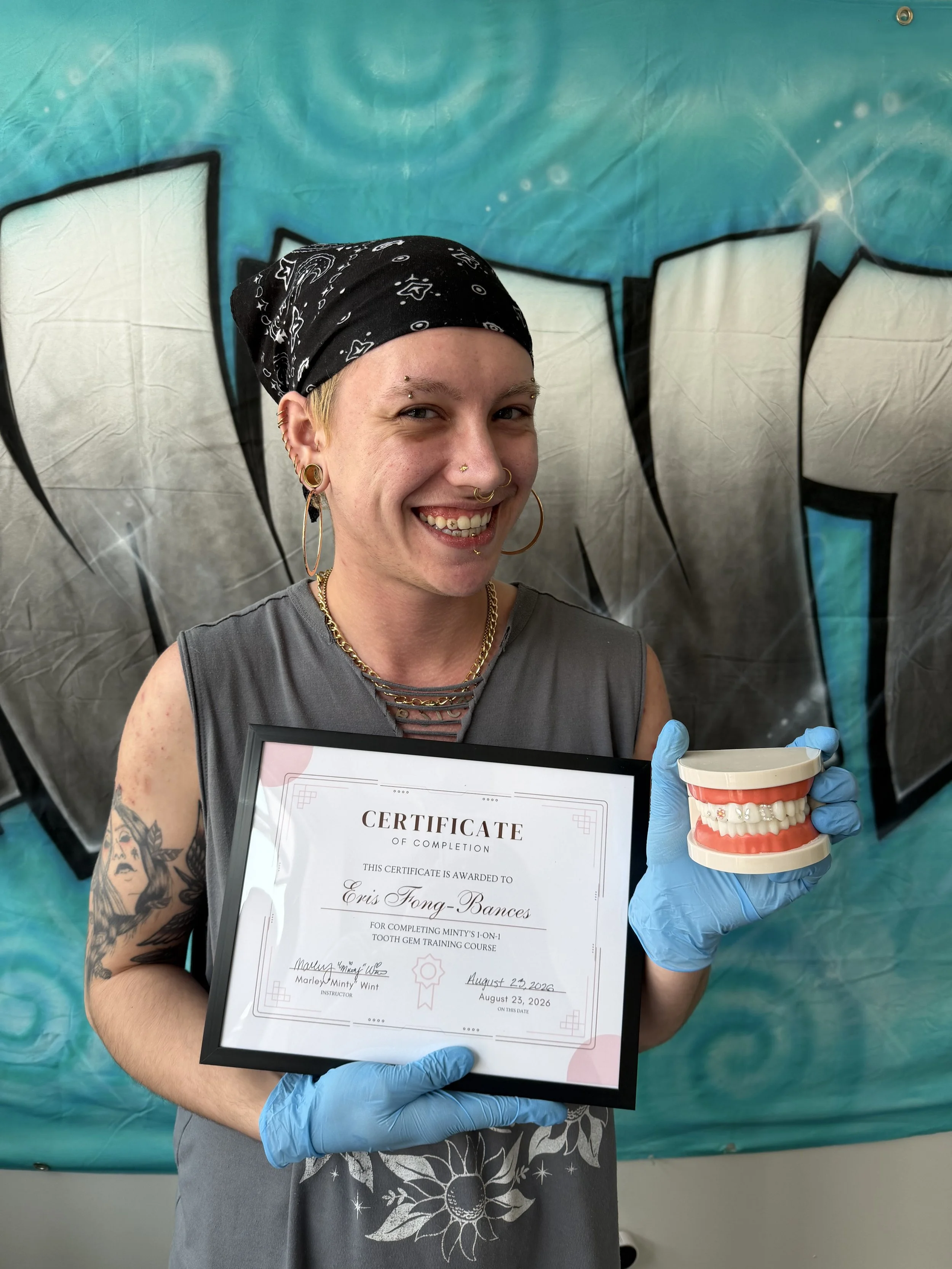 A white person smiling and holding a certificate and a dental model. They are wearing multiple facial piercings and earrings, a black bandana, a gray sleeveless shirt, and blue gloves. The background is a graffiti-style mural in blue and white.