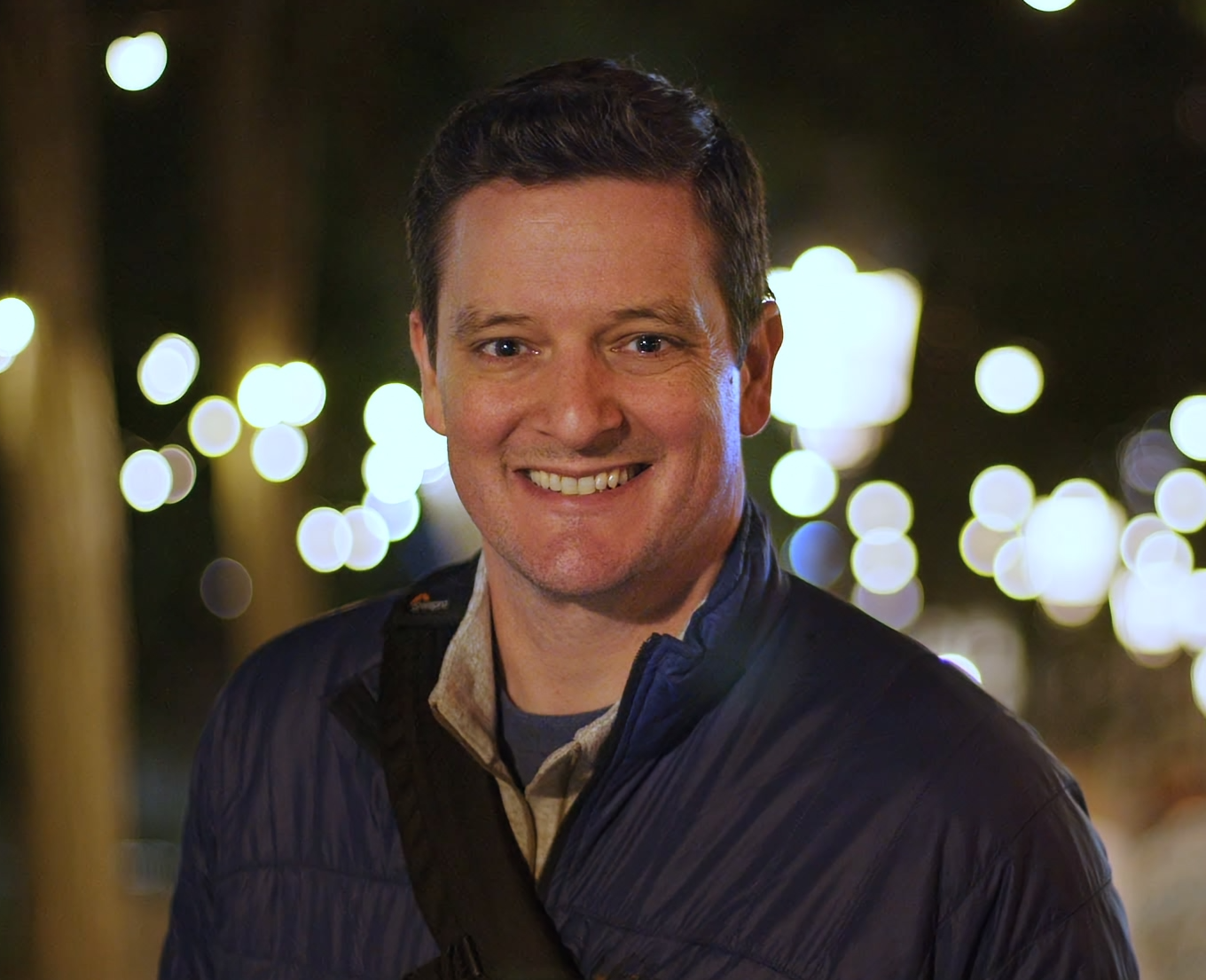 A cinematic shot of Matthew DeJohn, outdoors at night, with blurred city lights in the background.