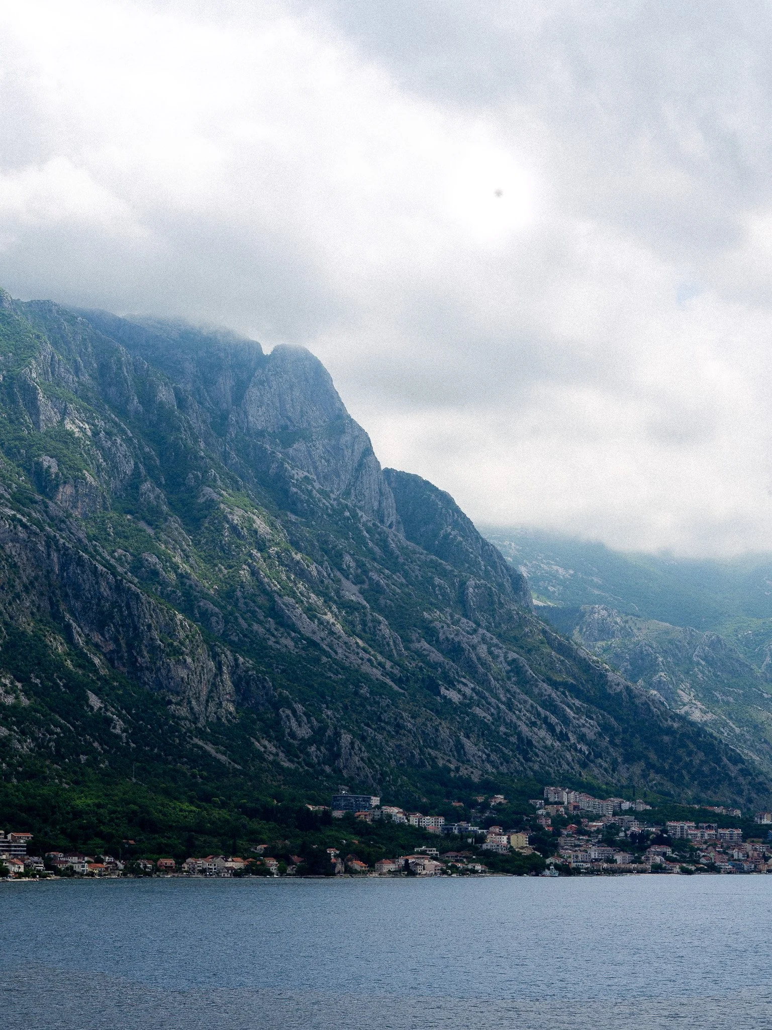 A scenic view of mountains with a small town at the base, next to a lake, under a cloudy sky. Montenegro.
