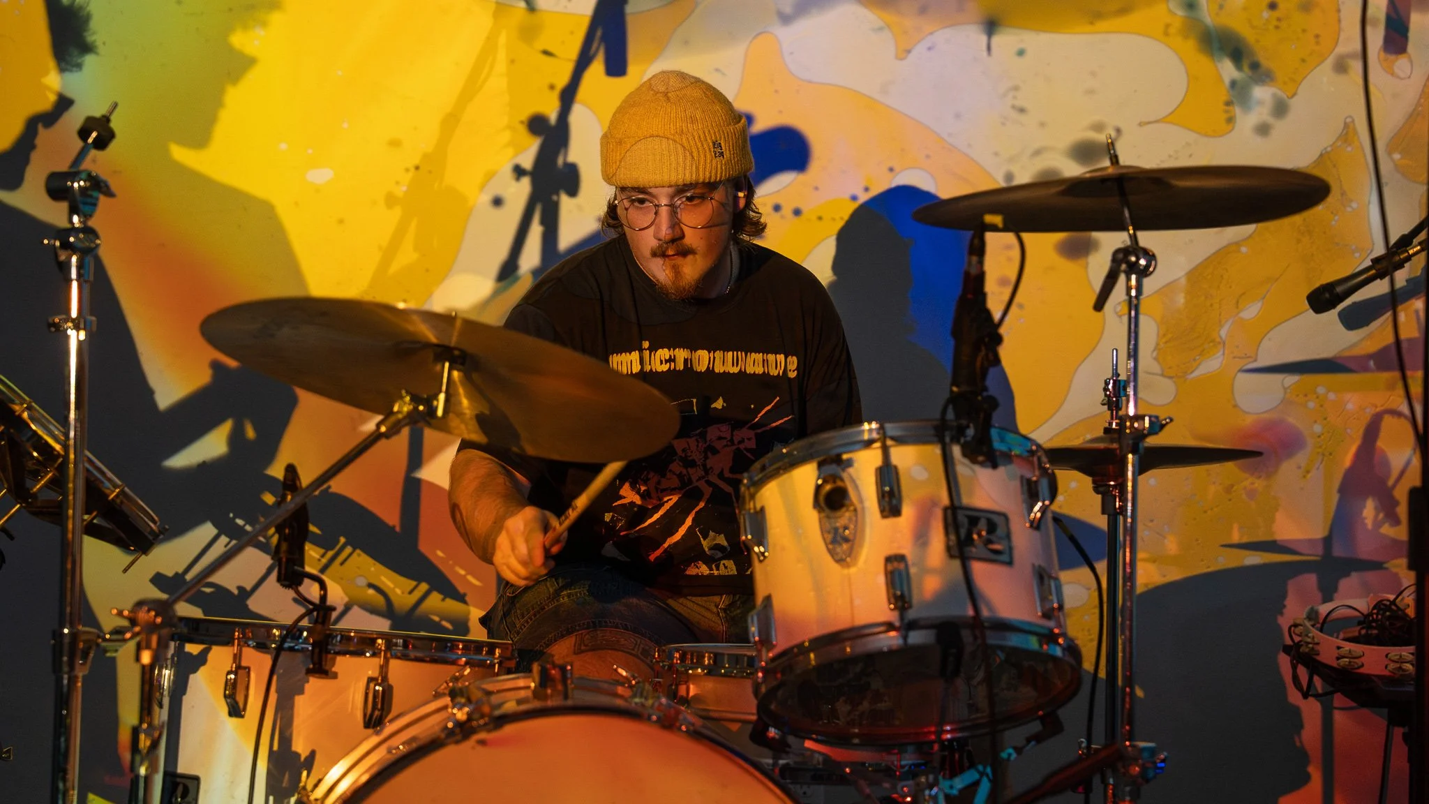 A man wearing glasses and a yellow knit cap playing a drum set on stage with colorful abstract background projection.