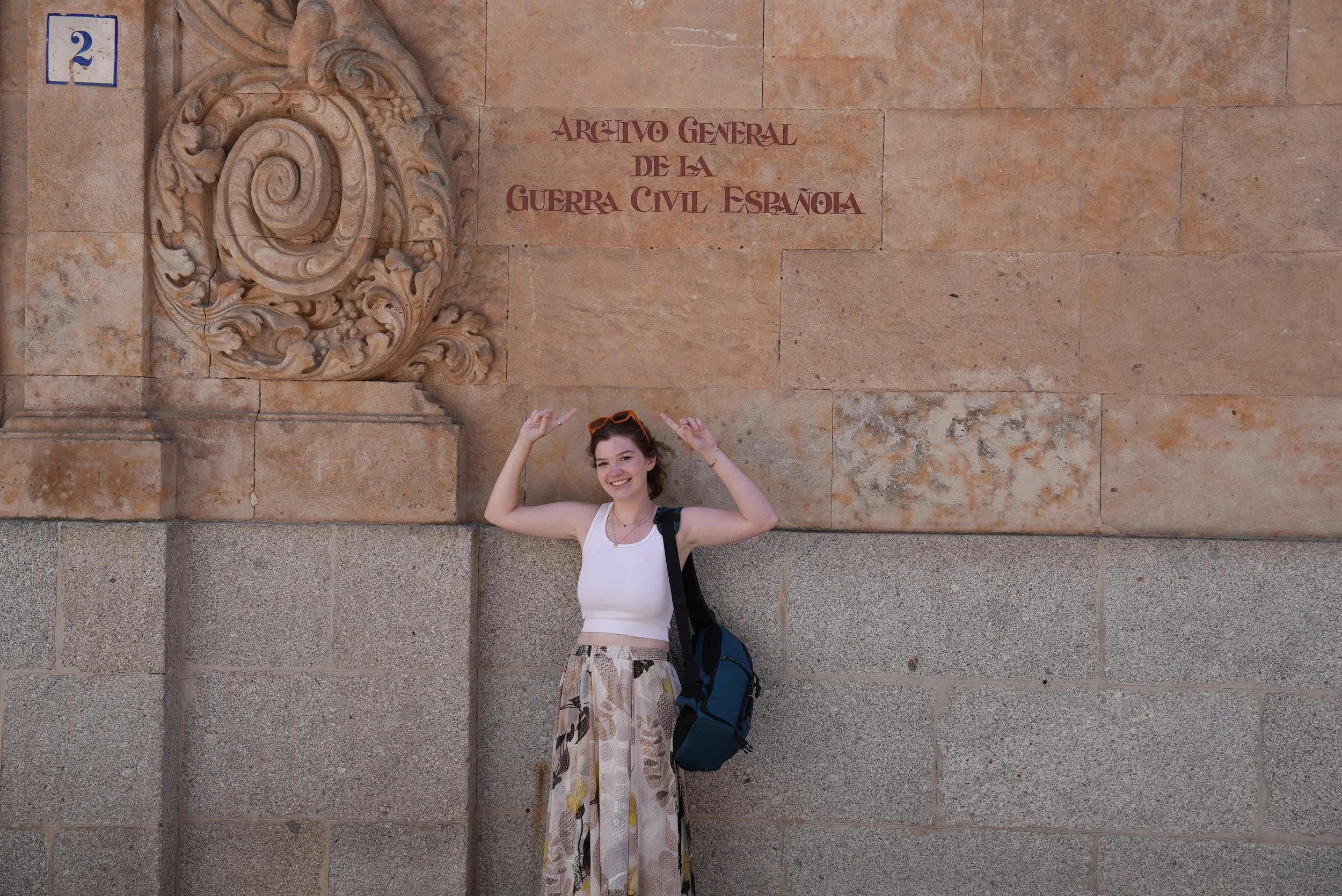 A woman smiling and pointing up with both hands stands in front of a beige stone wall. There is a decorative stone carving to the left and a sign that reads 'Archivo General de la Guerra Civil Española' above her head. She wears a white sleeveless top, patterned wide-leg pants, sunglasses atop her head, and carries a backpack.