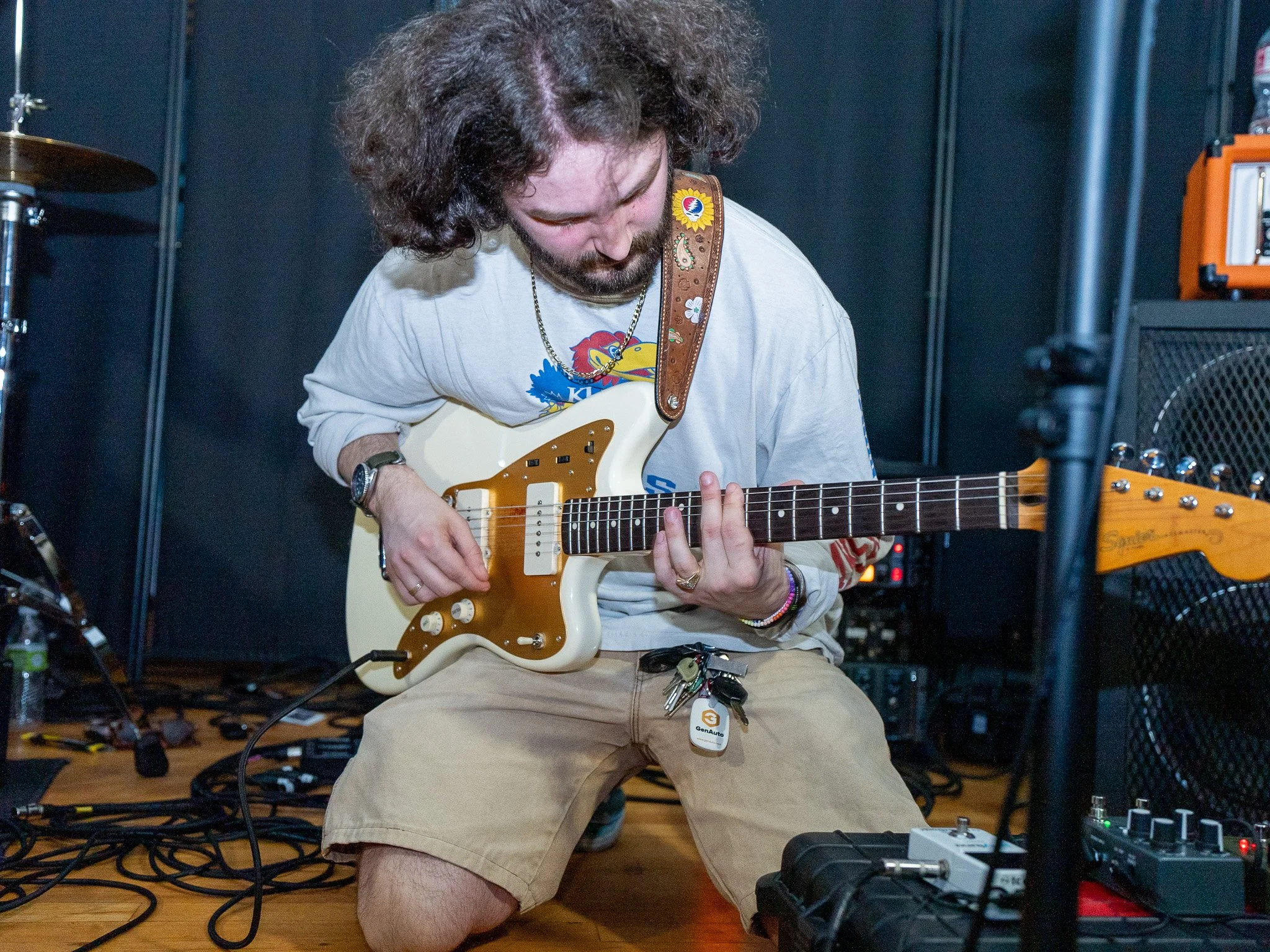 A man with curly hair and a beard, wearing a beige Kansas Jayhawks T-shirt and khaki shorts, is sitting on the floor playing an electric guitar. He has a rainbow-colored bracelet on his right wrist and a keychain attached to his shorts. The backgroun