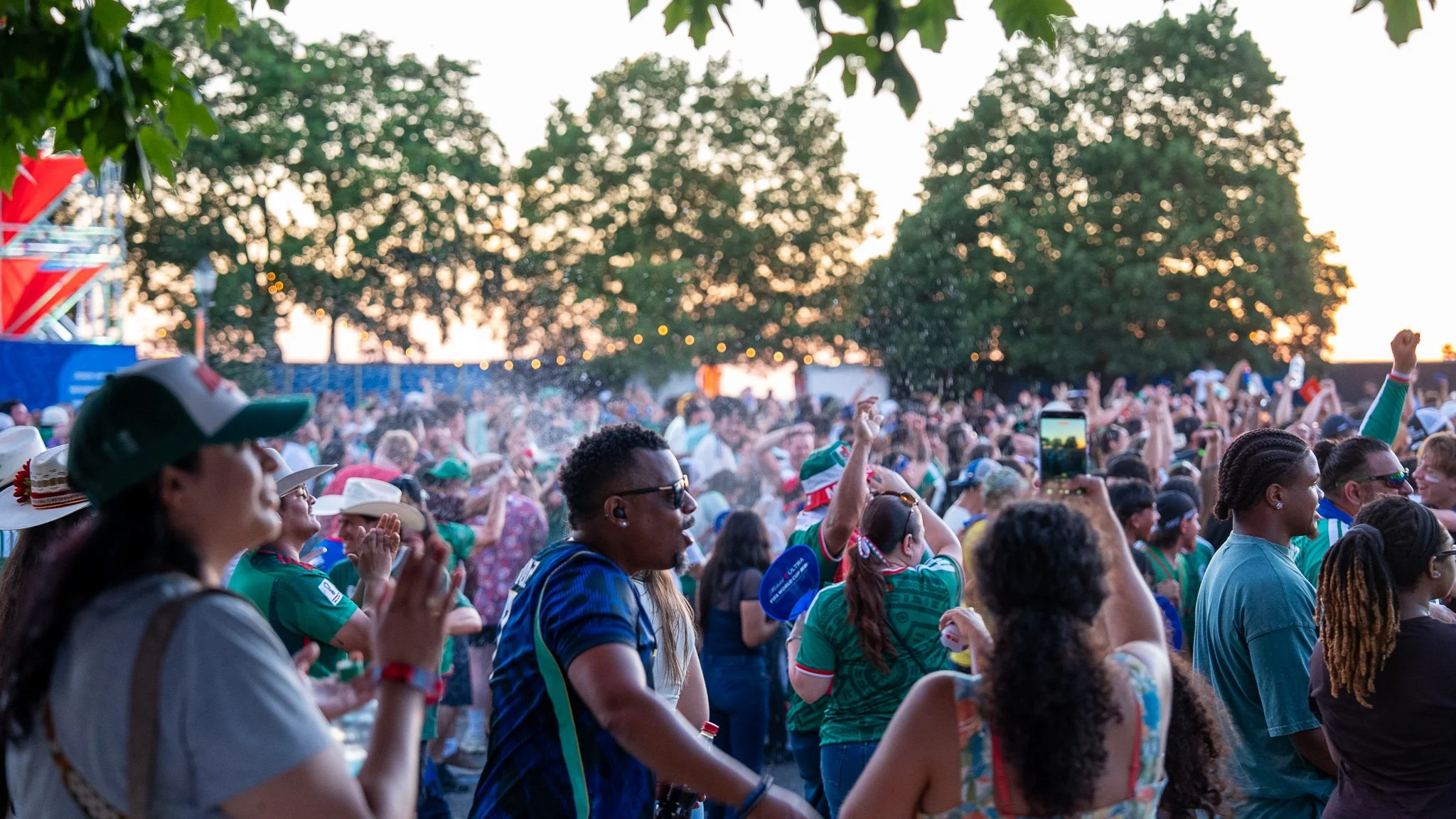 Crowd of people outdoors, some wearing sports jerseys and hats, watching an event during sunset with large trees in the background. FIFA fan fest.