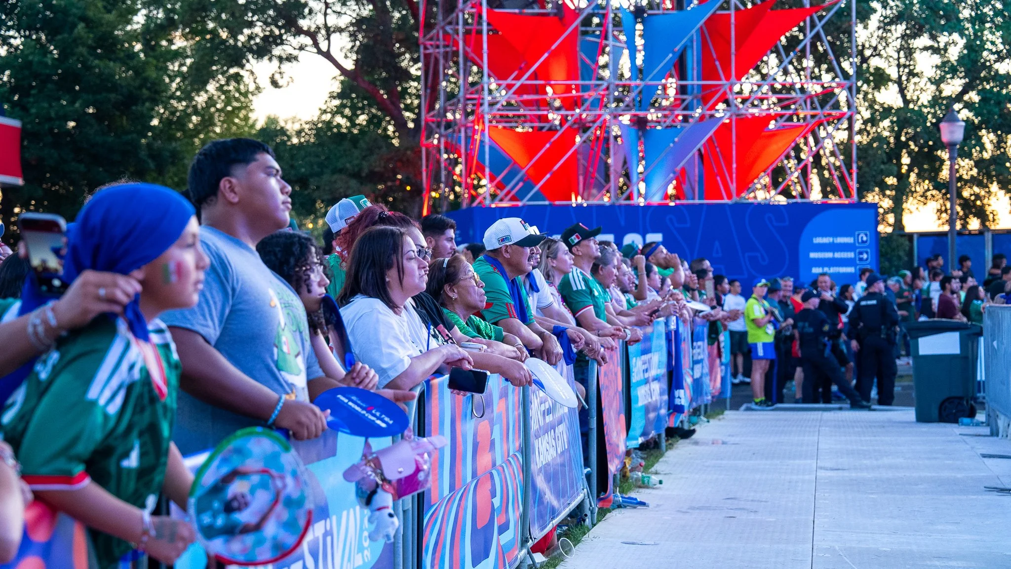 Crowd of people watching an outdoor event at sunset, with a colorful stage in the background.