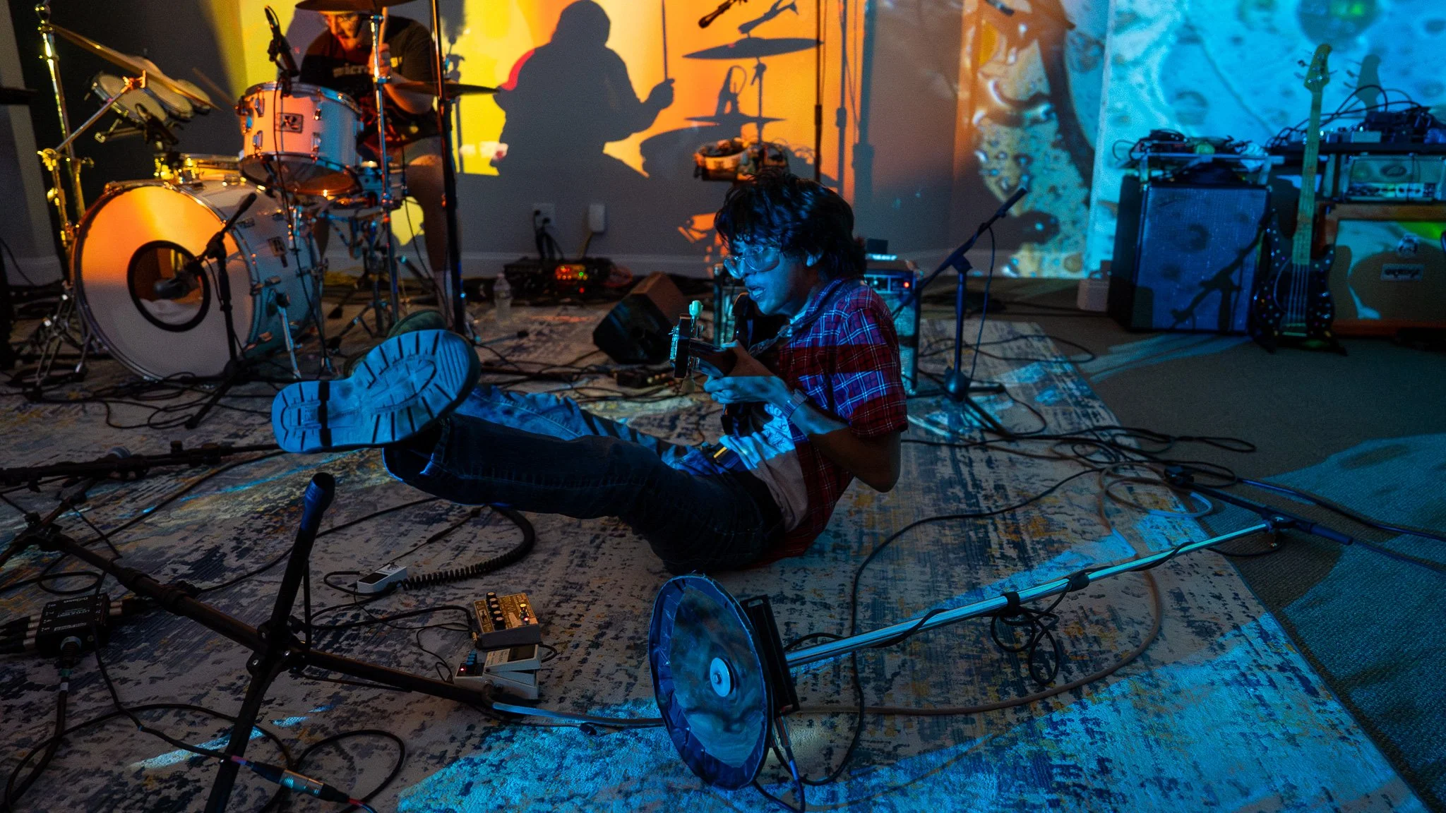 A musician sitting on the floor surrounded by musical instruments and equipment, playing an electric guitar with a colorful background and shadows of other band members projected on the wall.