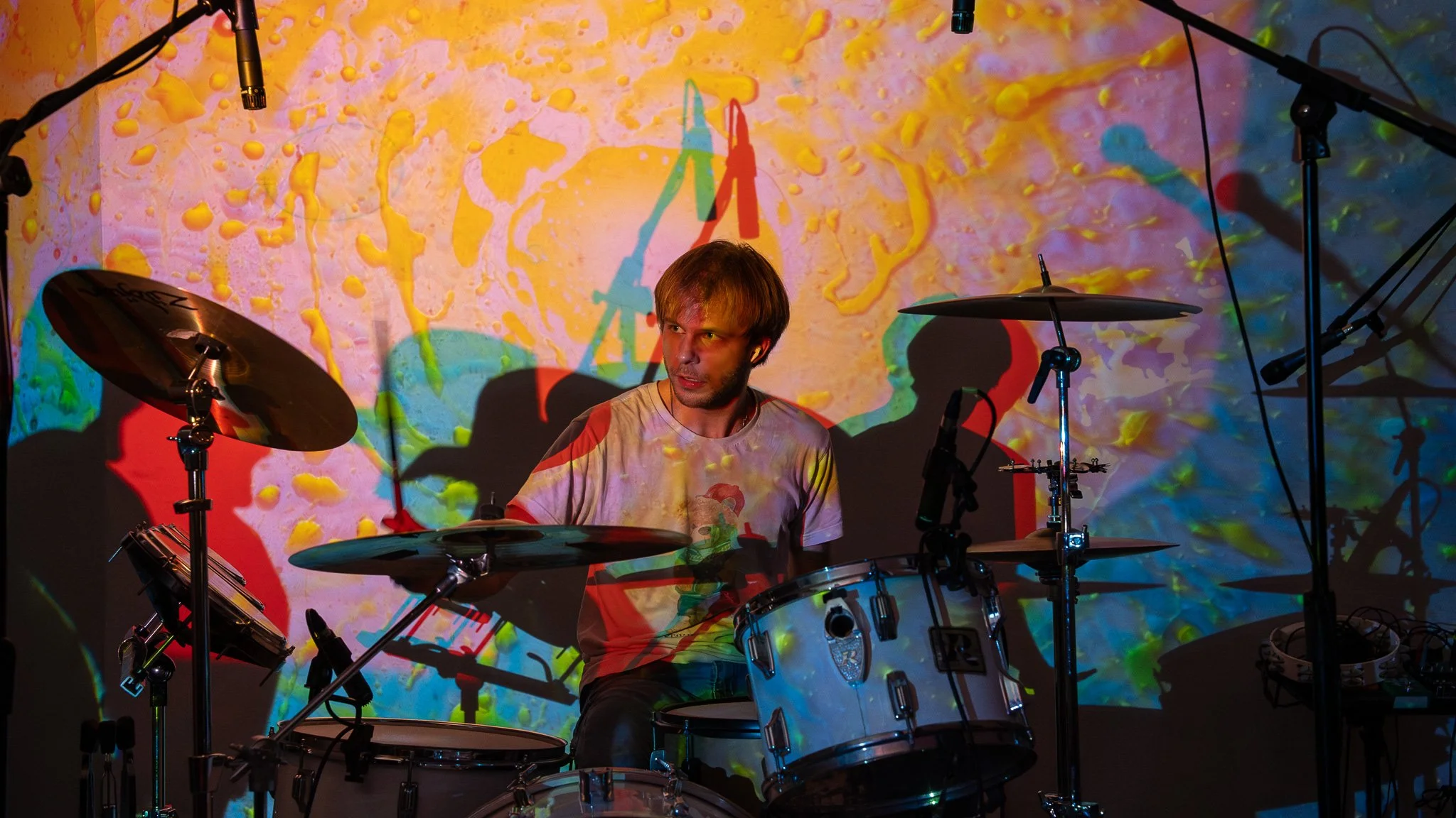 A man playing drums in front of a colorful, abstract projection with yellow, pink, blue, and green splashes.