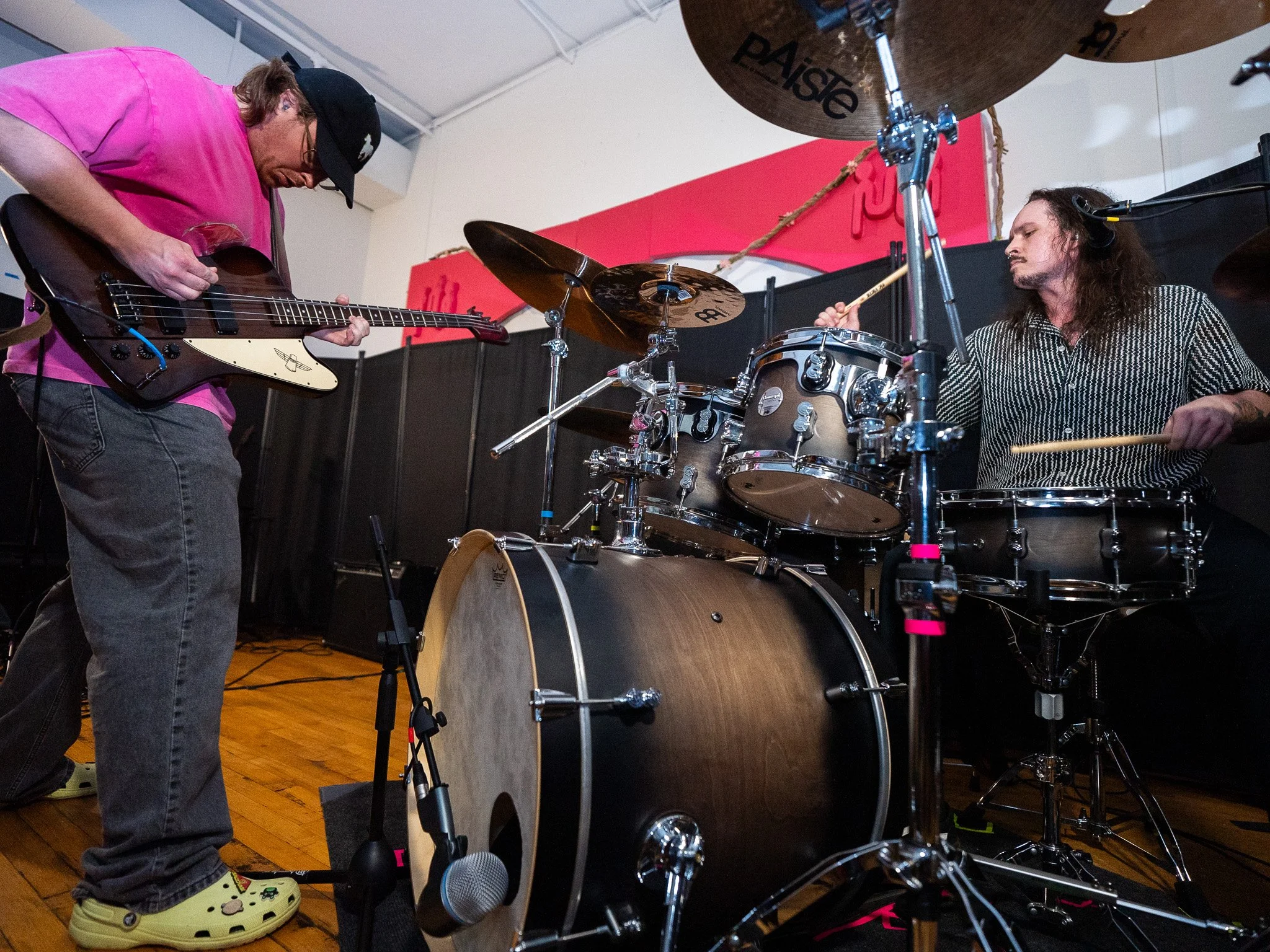 Two musicians performing with instruments: one person playing an electric bass guitar and another playing a drum set. The bassist is wearing a pink shirt, gray pants, a black cap, and Crocs. The drummer has long hair, a tattoo on the left arm, and is