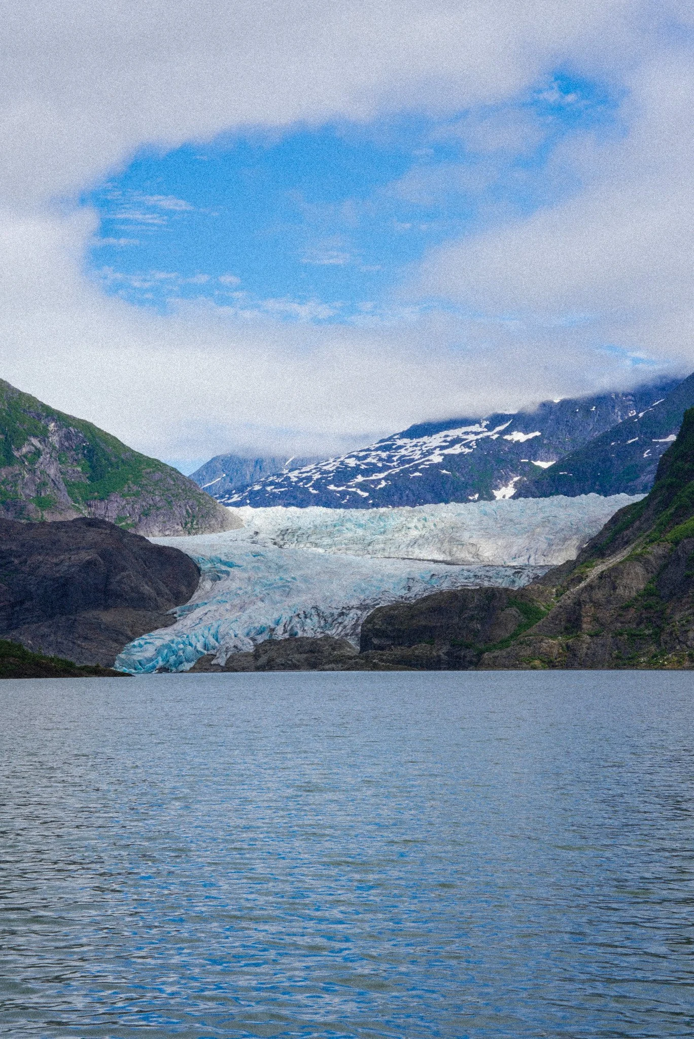 A scenic view of a glacier flowing from snow-capped mountains into a body of water, with partly cloudy sky above. Alaskan glacier