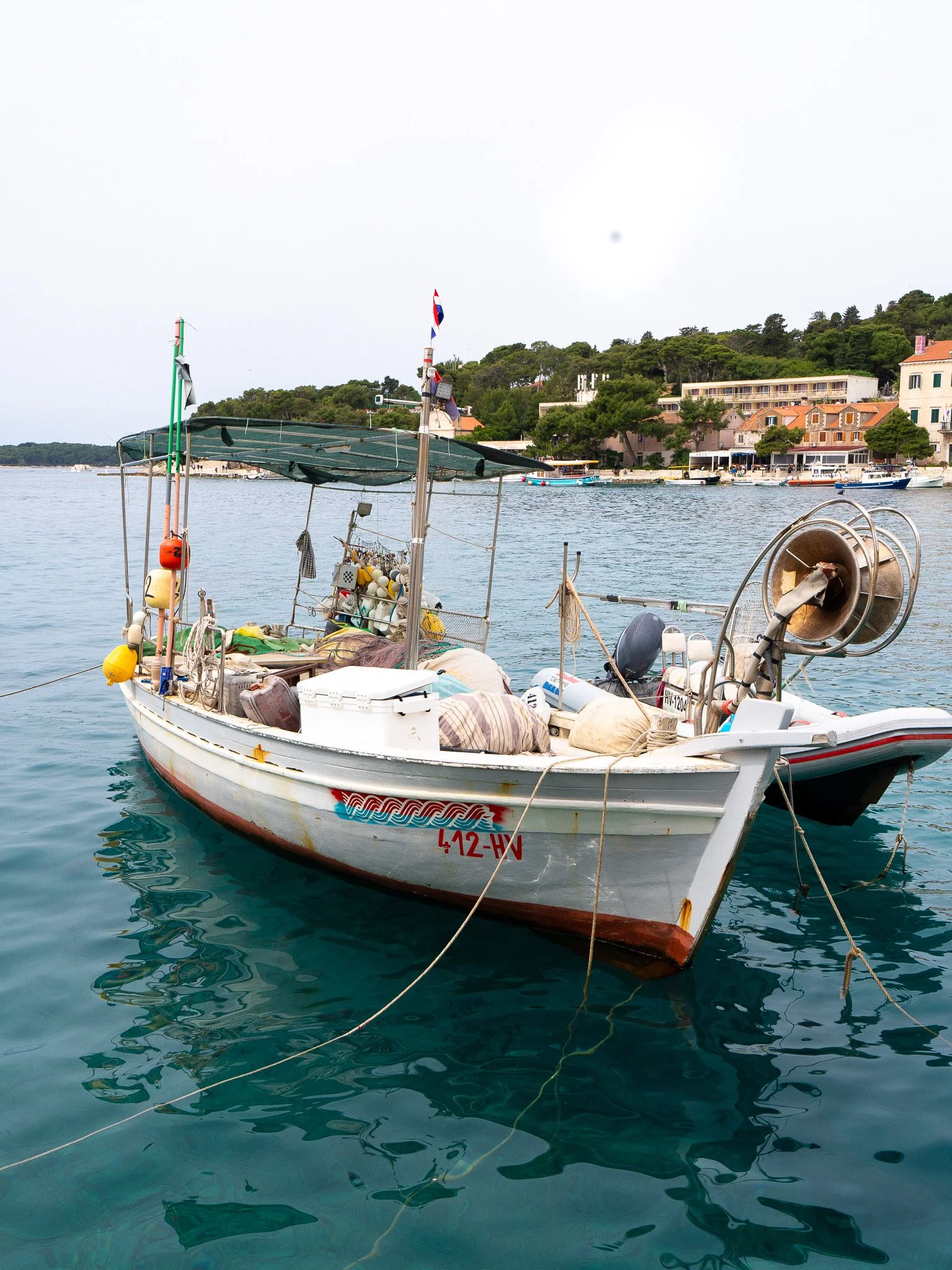 A small white boat with equipment and supplies on board, docked in a harbor with water, buildings, and trees in the background. Croatia.