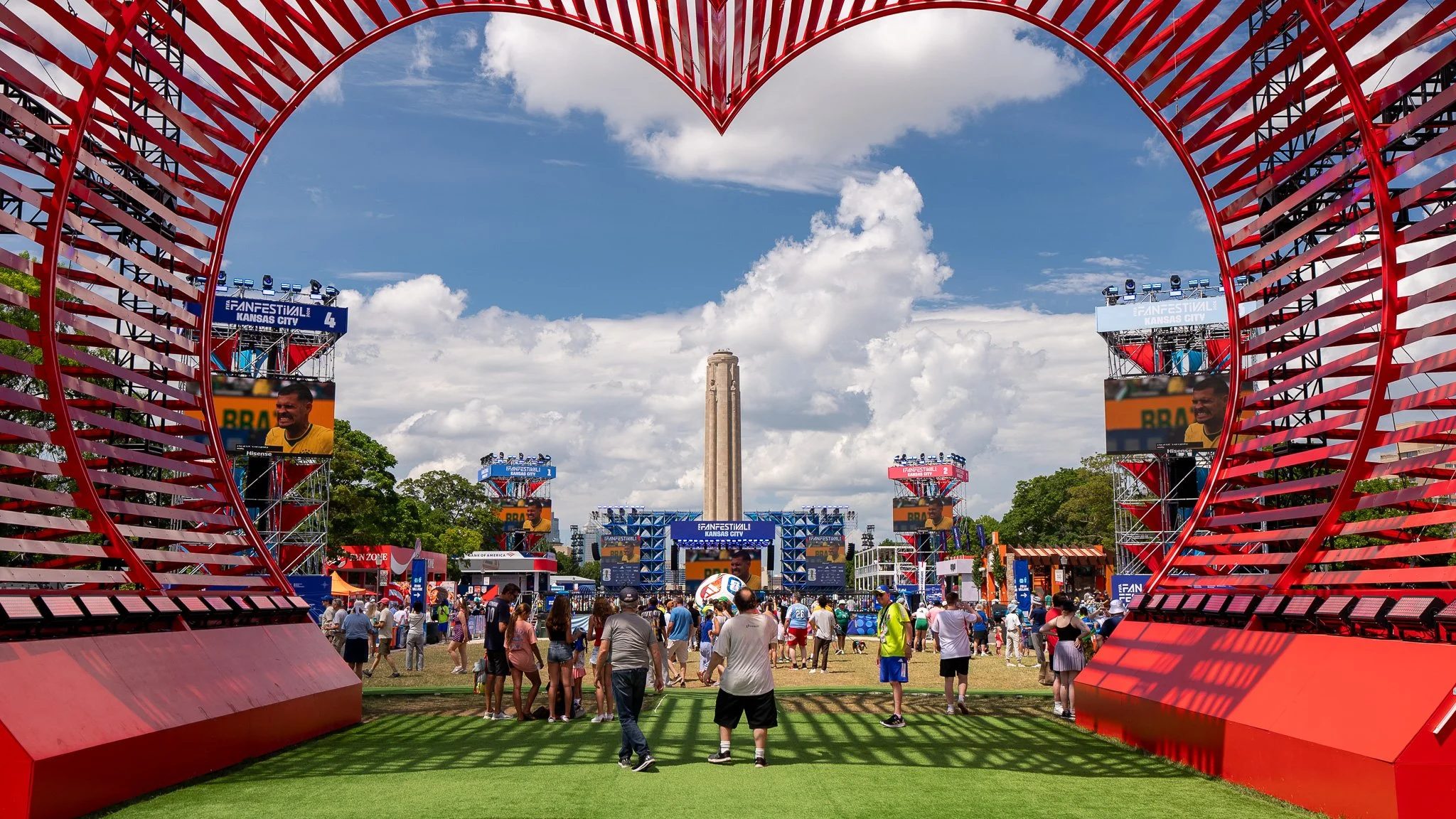 Crowd attending a large outdoor event with multiple screens, colorful banners, and a large stage, framed by a red metal arch structure under a partly cloudy sky.