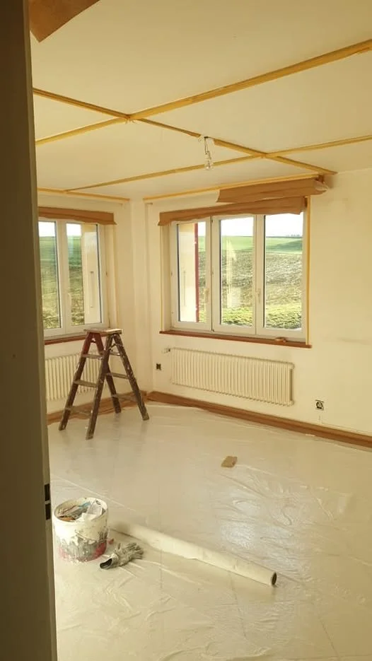 Salle en rénovation avec fenêtres, radiateurs, un escabeau en bois, et des outils de peinture sur le sol.