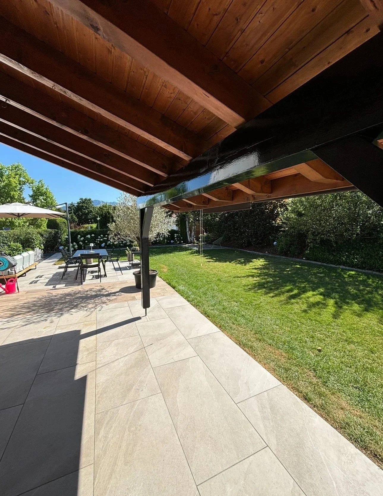 Patio en pierre avec mobilier de jardin comprenant une table, des chaises, un parasol, un lavabo extérieur et un espace vert avec des arbres et des buissons.