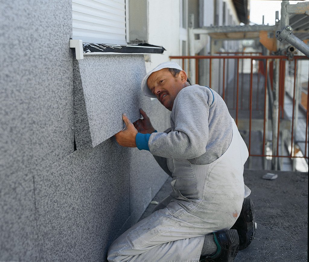 Un homme travaillant sur le revêtement extérieur d'un bâtiment, installant ou réparant un panneau en pierre ou en céramique.