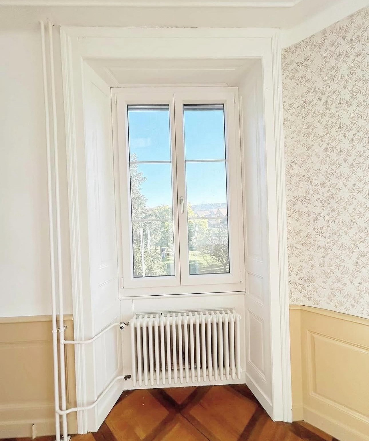 Une fenêtre blanche donnant sur un paysage extérieur avec des arbres et un ciel bleu, et un radiateur en dessous, dans une pièce avec des murs peints en beige et un papier peint à motifs floraux.