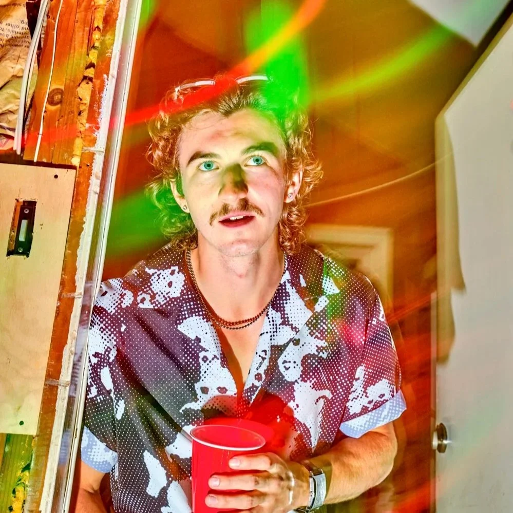 A man with curly hair and a mustache holding a red cup, standing in a room with wooden walls. There are colorful light streaks across the photo, creating a psychedelic effect.