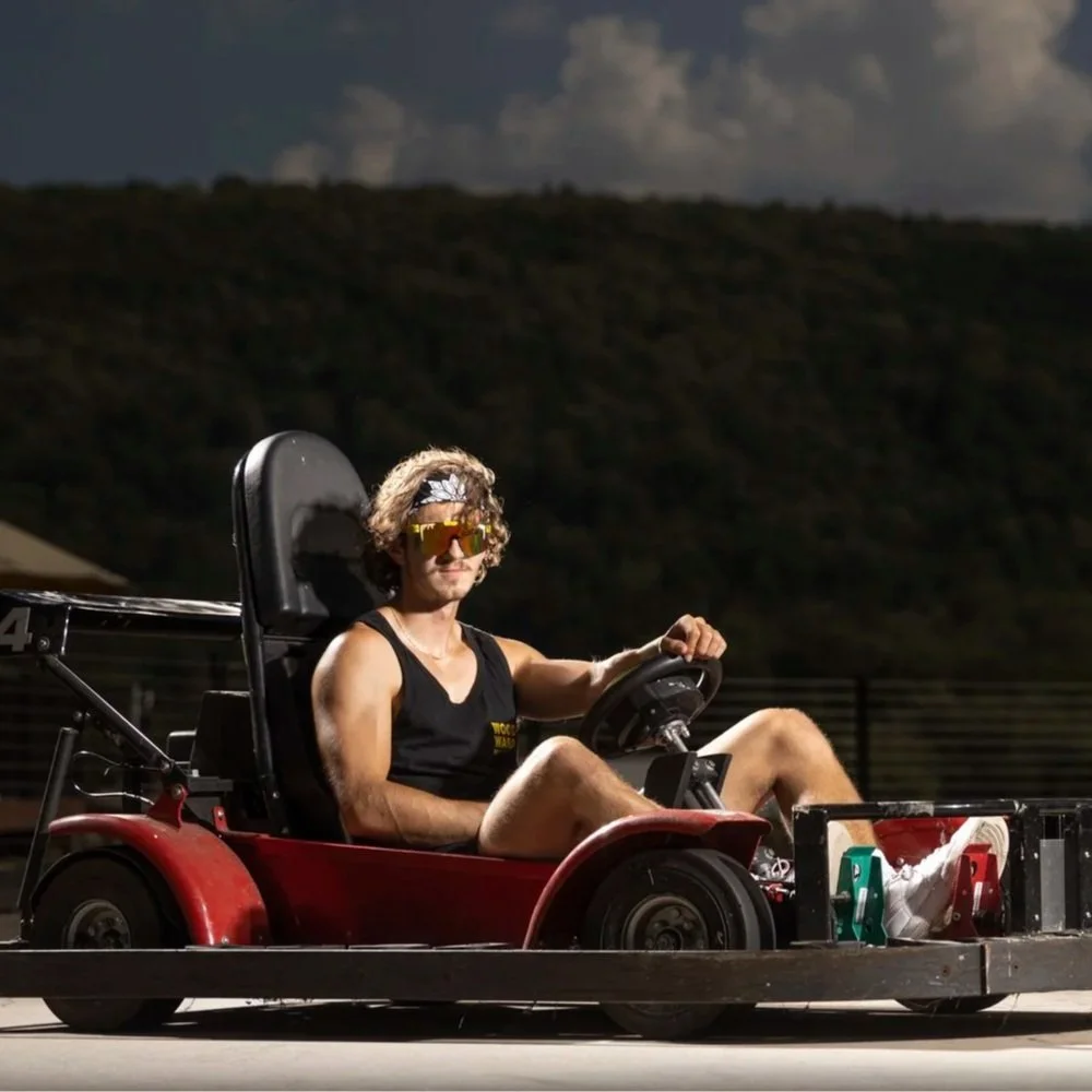 A young man with curly hair, sunglasses, and a bandana driving a small, red go-kart on a track at night, with a dark, hilly landscape in the background.