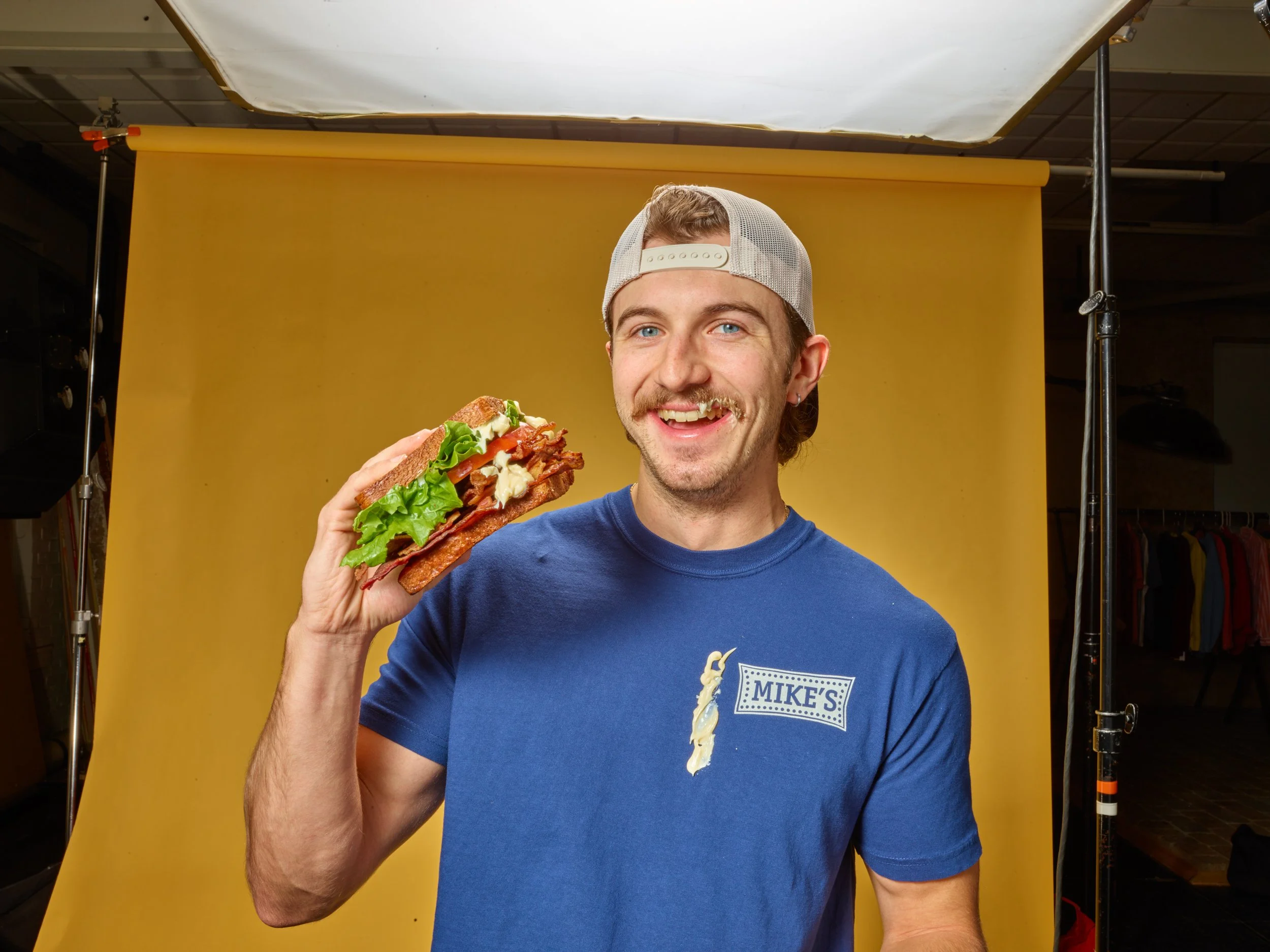 A young man wearing a backwards baseball cap and a blue T-shirt, smiling and holding a large sandwich with lettuce, tomato, bacon, and mayonnaise, in front of a yellow backdrop.