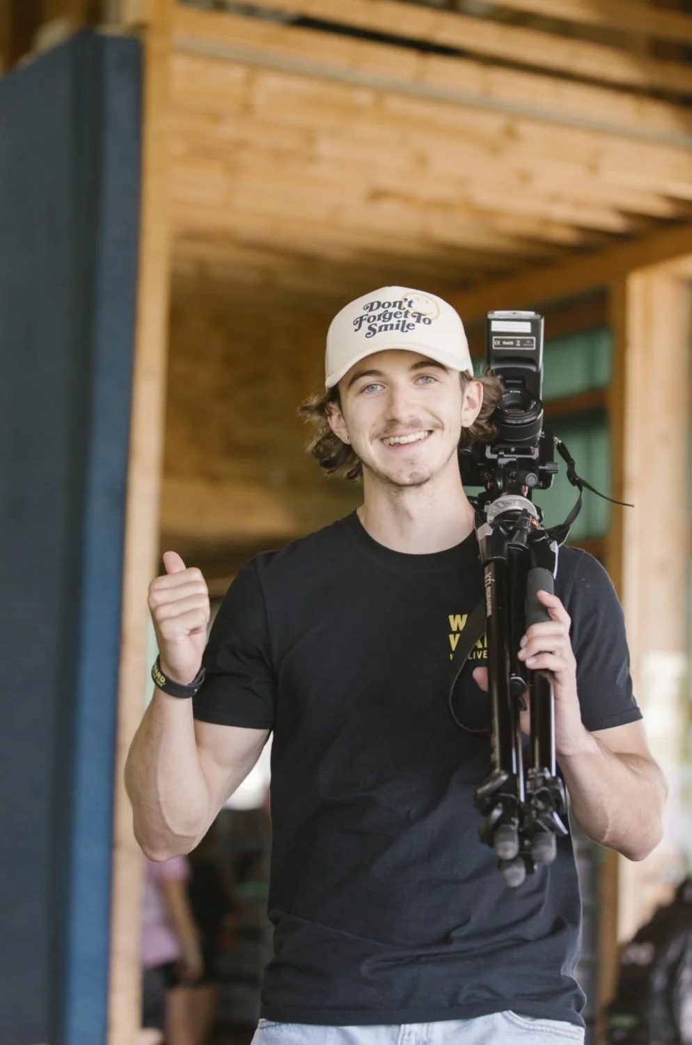 Young man smiling, wearing a white cap that says 'Don't Forget To Smile', holding a camera on a tripod over his shoulder, standing indoors with wooden walls.