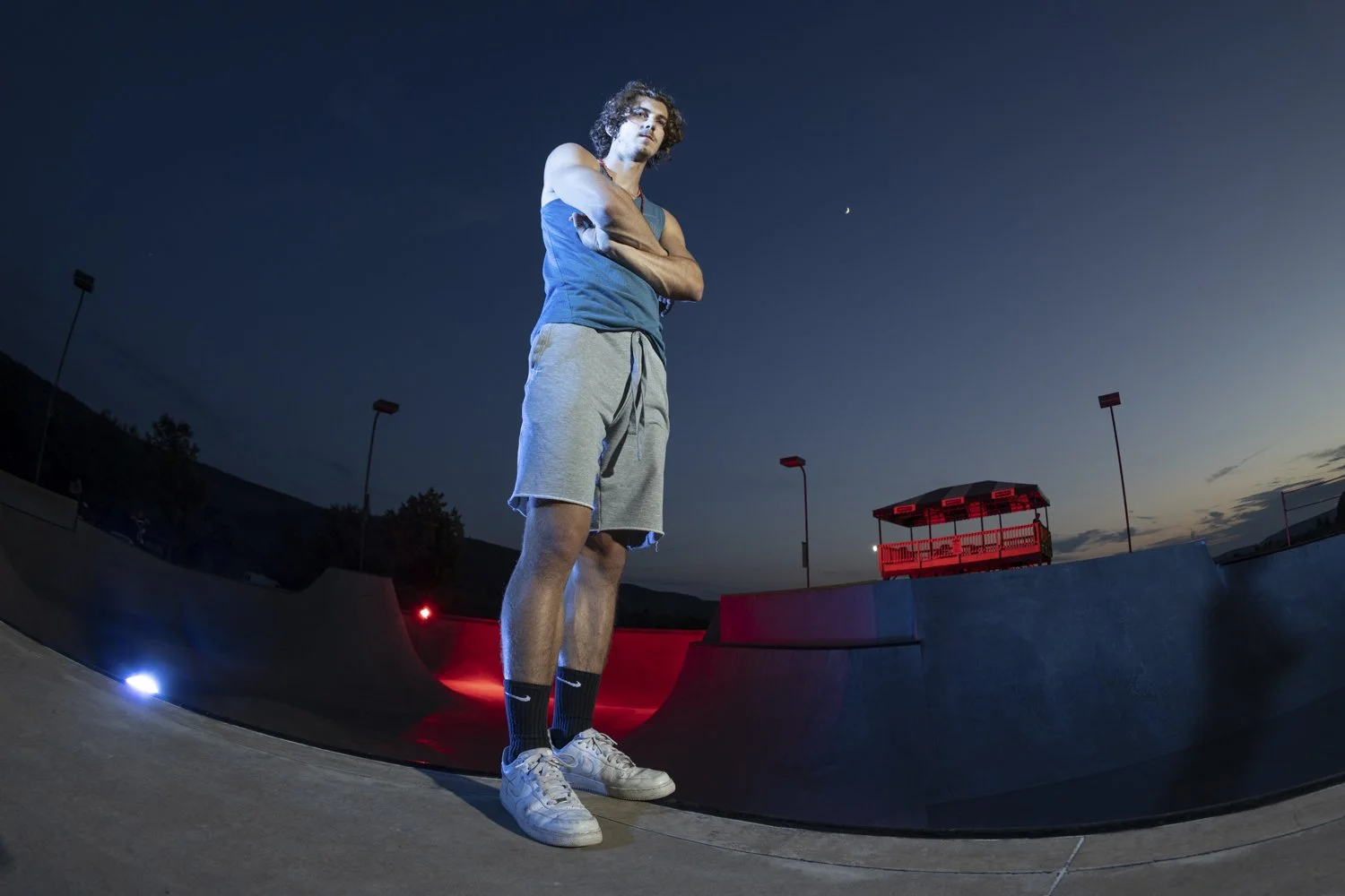 A young man with curly hair, wearing a blue tank top, gray shorts, black socks, and white sneakers, standing with arms crossed at a skatepark during dusk or night, with a dark sky, constellations, and a crescent moon visible in the background.