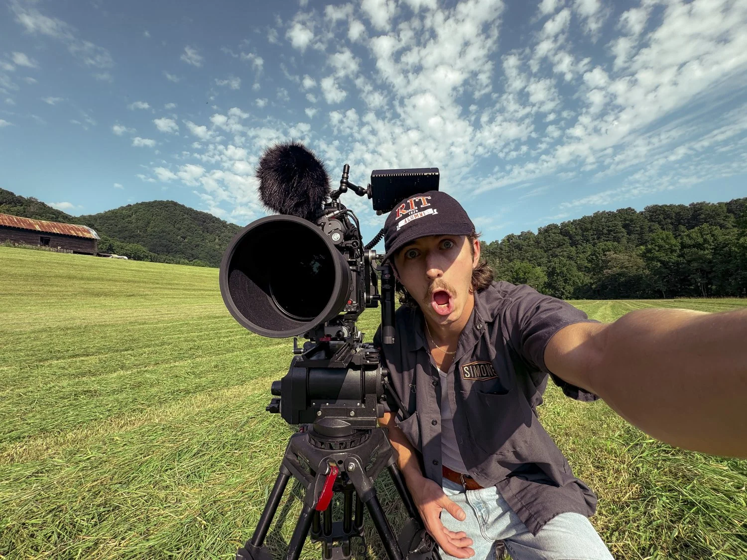 A man wearing a black cap and gray shirt taking a selfie with a professional video camera on a grassy field, with a background of trees, hills, an old barn, and blue sky with clouds.