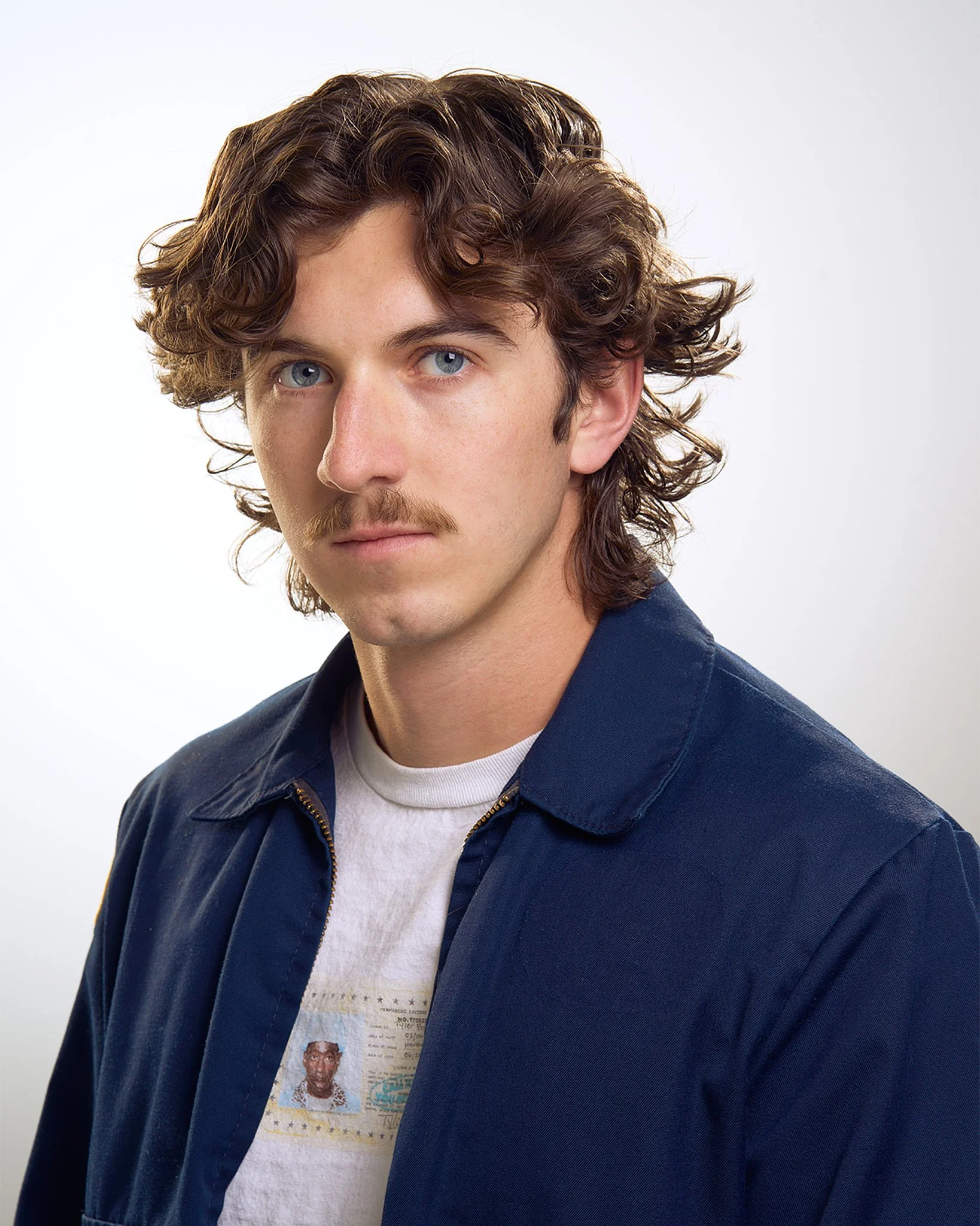 Close-up of a young man with curly brown hair, blue eyes, a mustache, wearing a navy blue jacket over a white T-shirt with an ID badge print, against a plain white background.