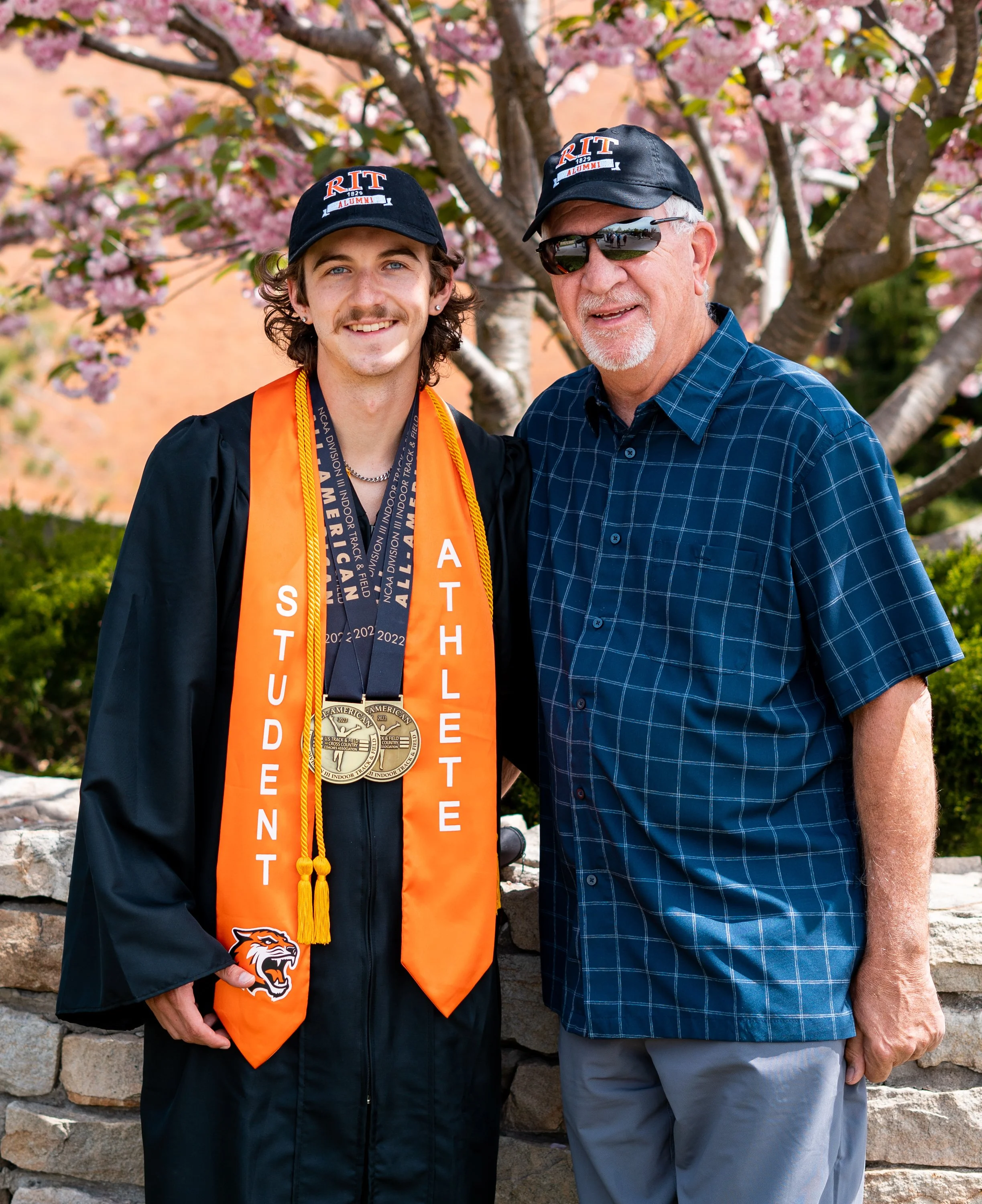 A young man in a graduation gown and cap with the university logo, and an older man in a plaid shirt and hat with the university logo, standing outdoors in front of a flowering tree, both smiling for the photo.