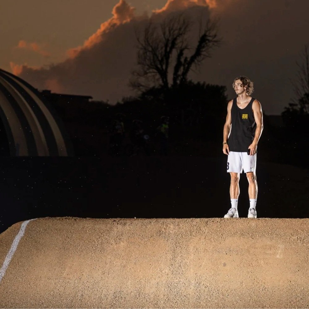 A young man with curly hair, glasses, and a sleeveless basketball jersey and shorts stands on a dirt surface at sunset, with dark clouds and trees in the background.
