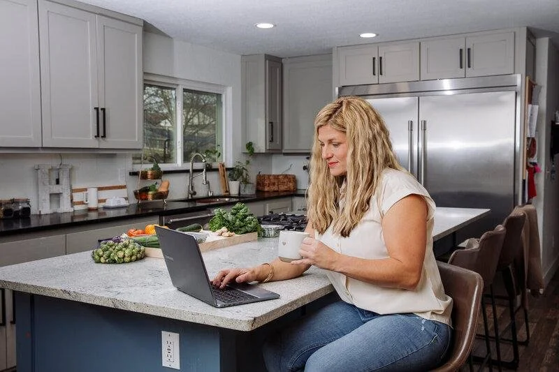 Woman sitting at kitchen island using a laptop and holding a mug.