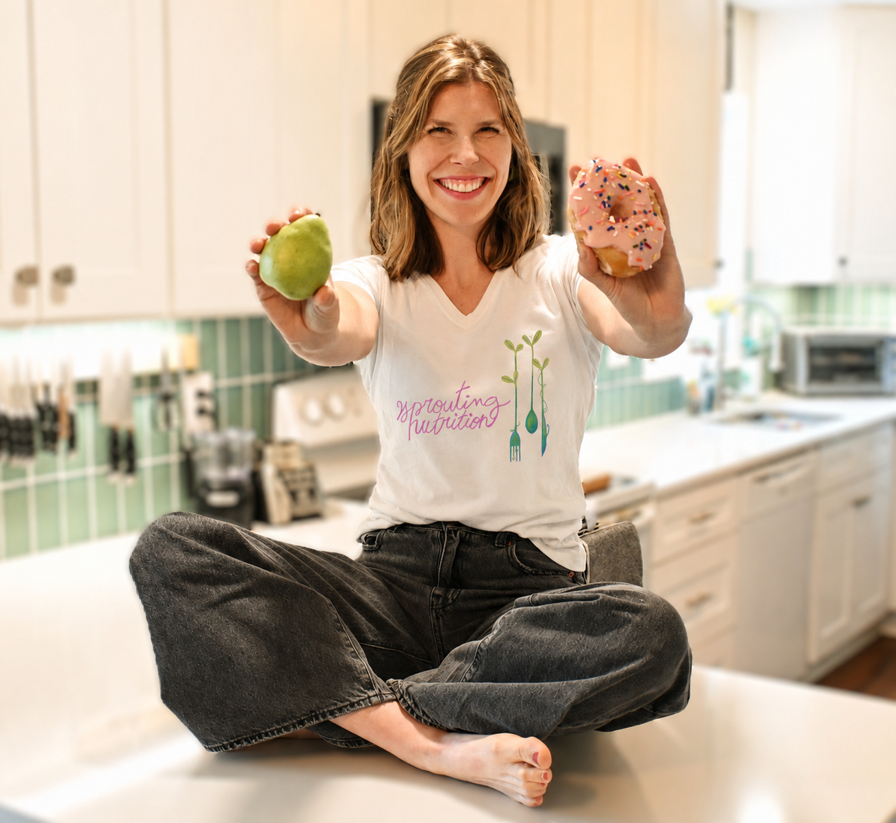 A woman sitting cross-legged on a kitchen countertop, smiling and holding a green apple in her left hand and a pink frosted donut with sprinkles in her right hand.