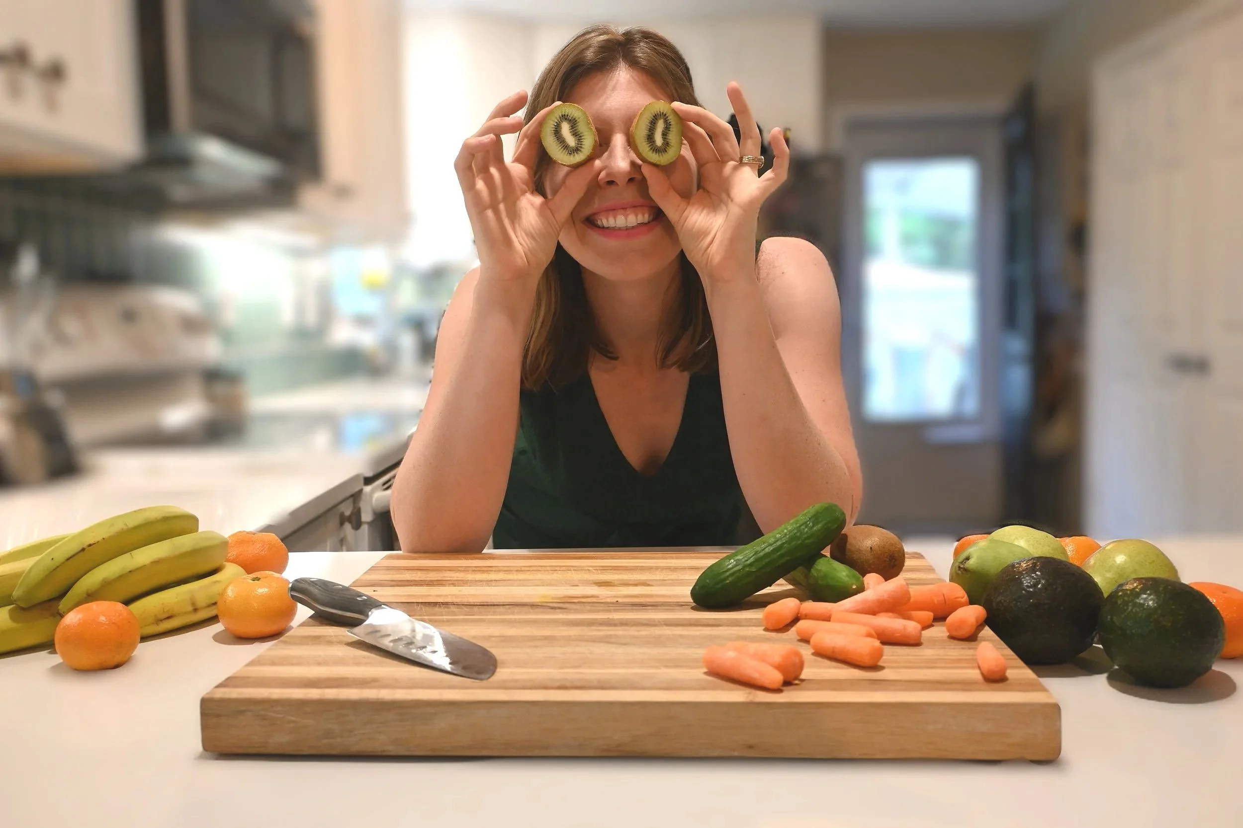 Woman with slices of kiwi over her eyes, smiling in a kitchen, surrounded by various fruits and vegetables including bananas, oranges, cucumbers, carrots, and avocados on a wooden cutting board.