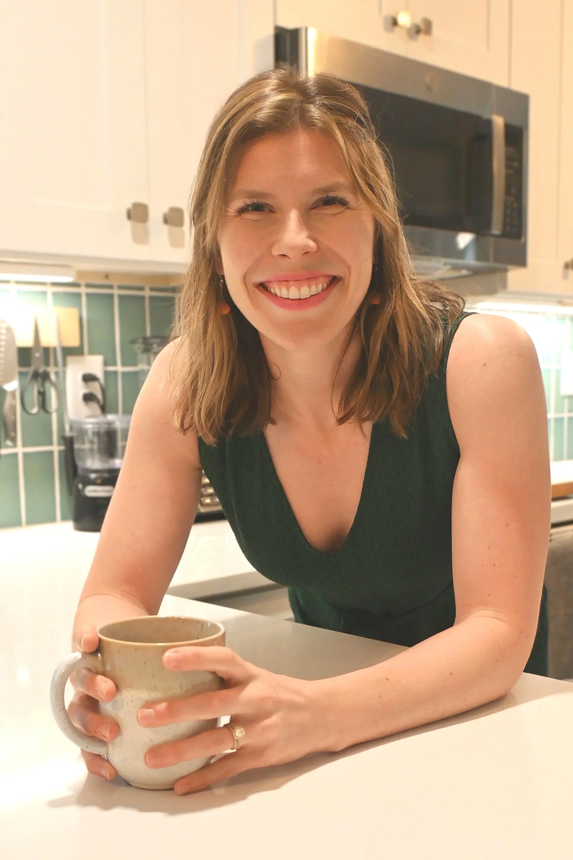 A woman smiling and holding a coffee mug in a kitchen. She has shoulder-length brown hair, is wearing a black sleeveless top, and appears to be sitting at a kitchen counter.